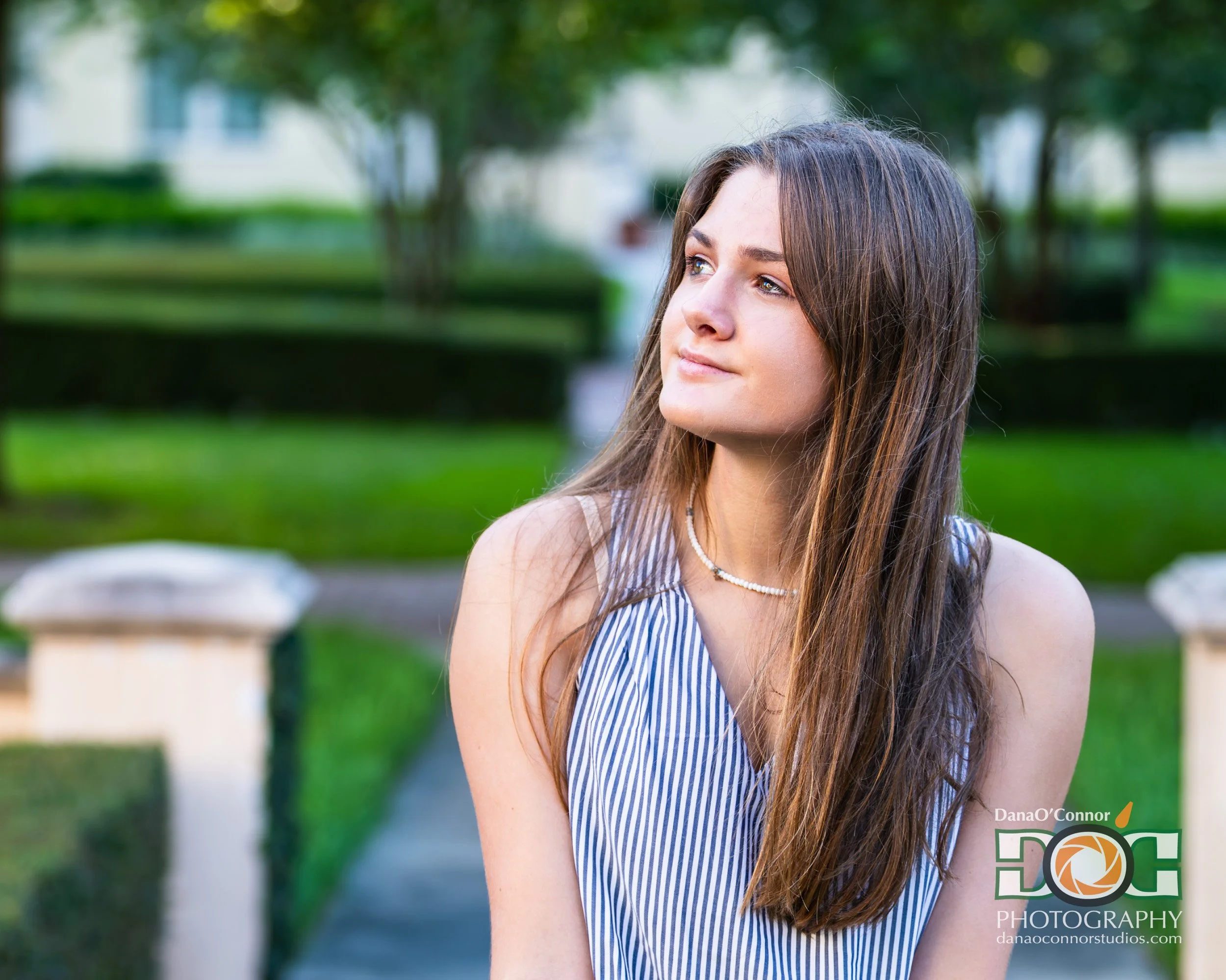 Young woman with long brown hair and blue eyes, wearing a blue and white striped sleeveless top, standing outdoors with a blurred green park background.