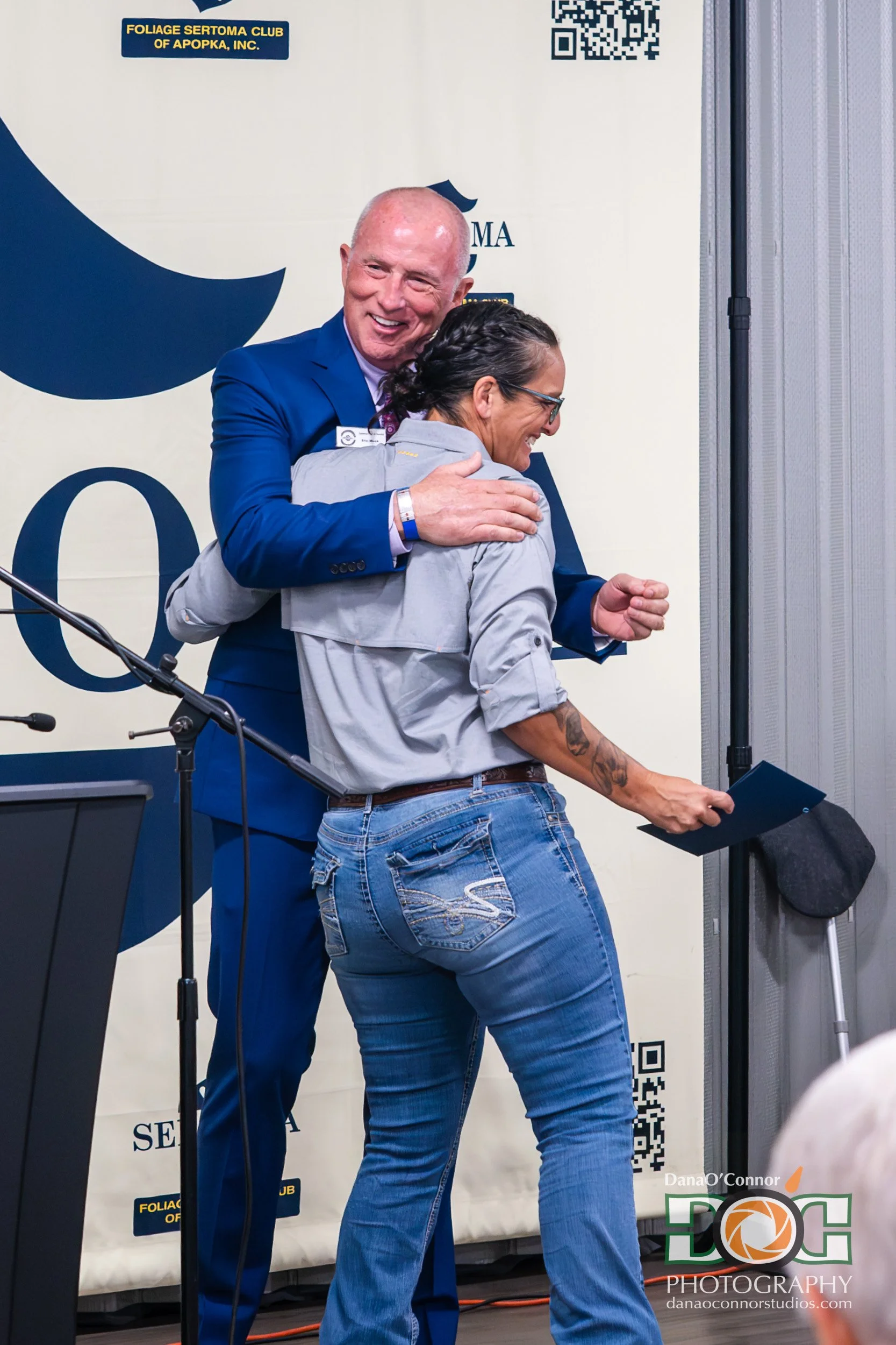 A man in a blue suit hugging a woman in sunglasses and a gray jacket at an indoor event, with a backdrop displaying logos and QR codes.