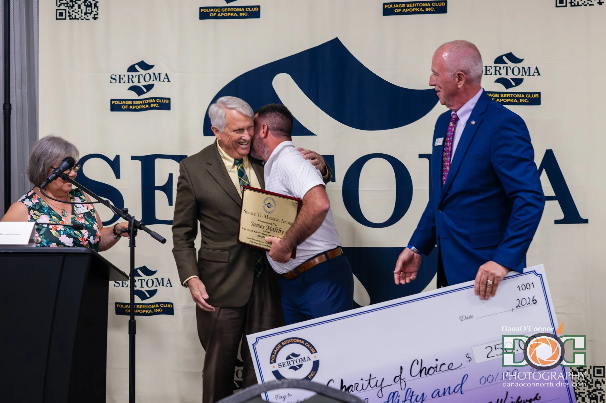 A man in a white shirt and brown pants receiving an award from a man in a gray suit at a ceremony. Two large checks are partially visible, and a woman with glasses and a floral dress stands at a podium with a microphone. A man in a blue suit and pink