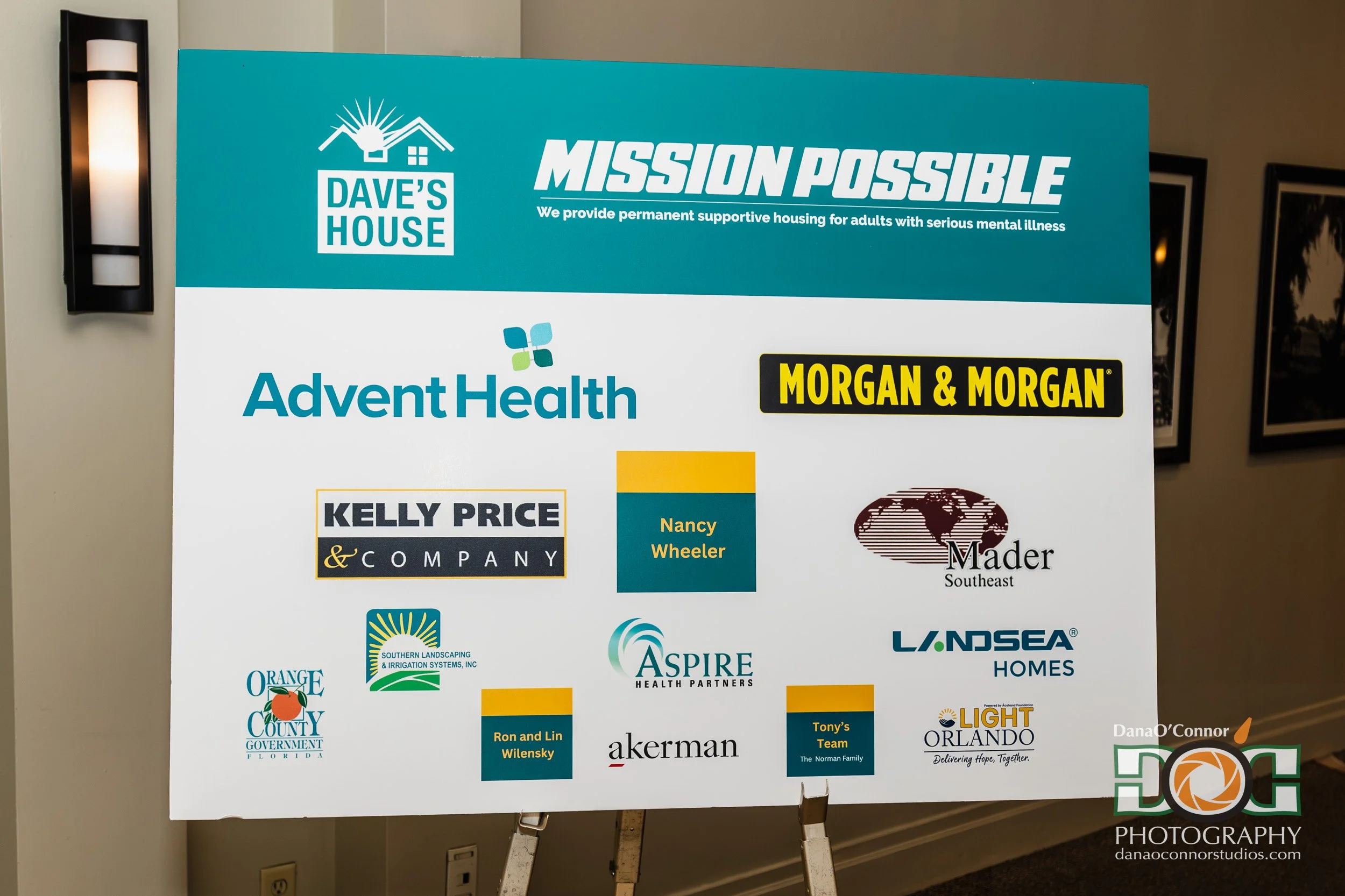 A large poster with various sponsor logos, including Dave's House, Advent Health, Morgan & Morgan, Kelly Price, and others, displayed on an easel.