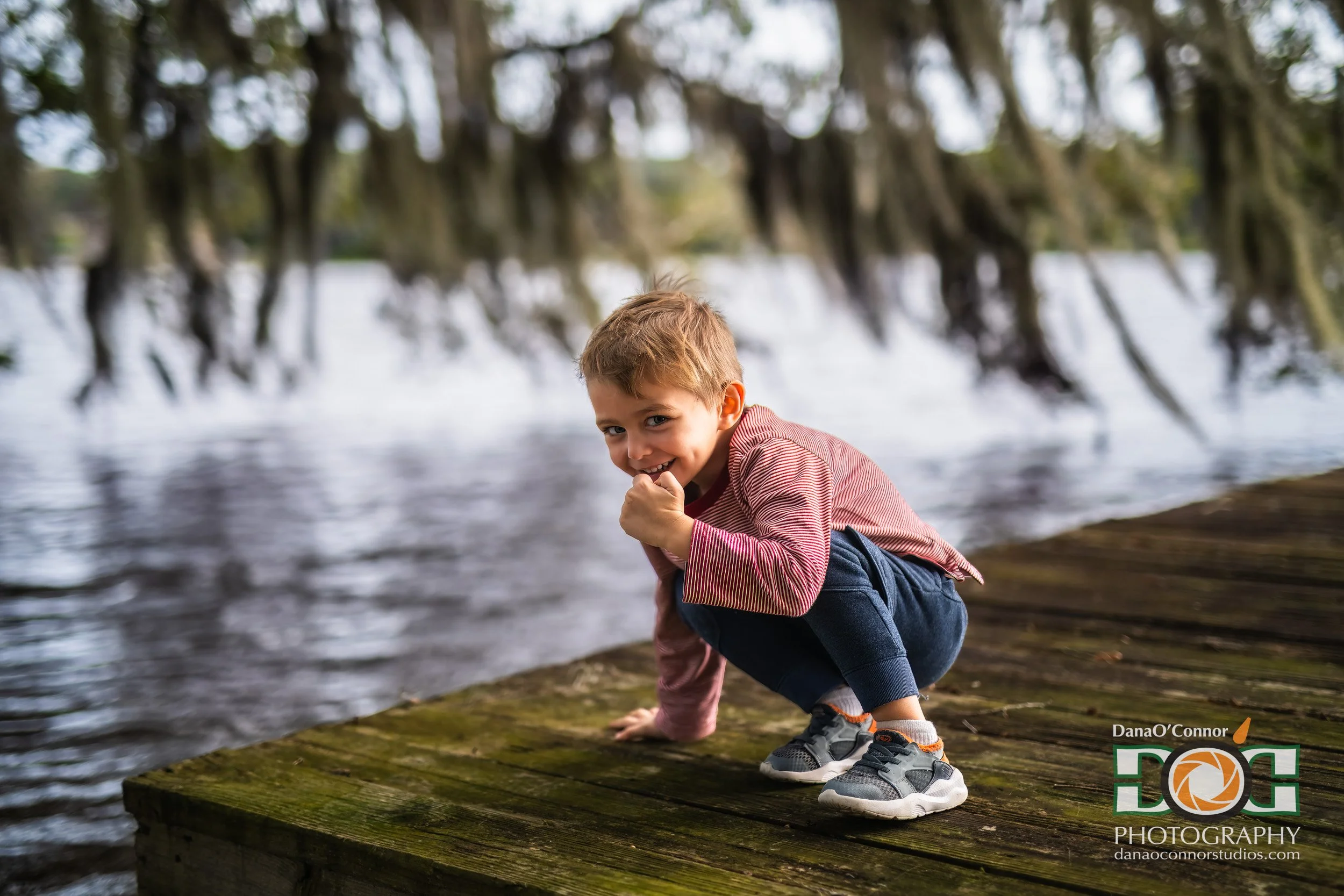 A young boy in a striped shirt and jeans crouches on a mossy dock near a body of water, smiling at the camera with trees in the background.