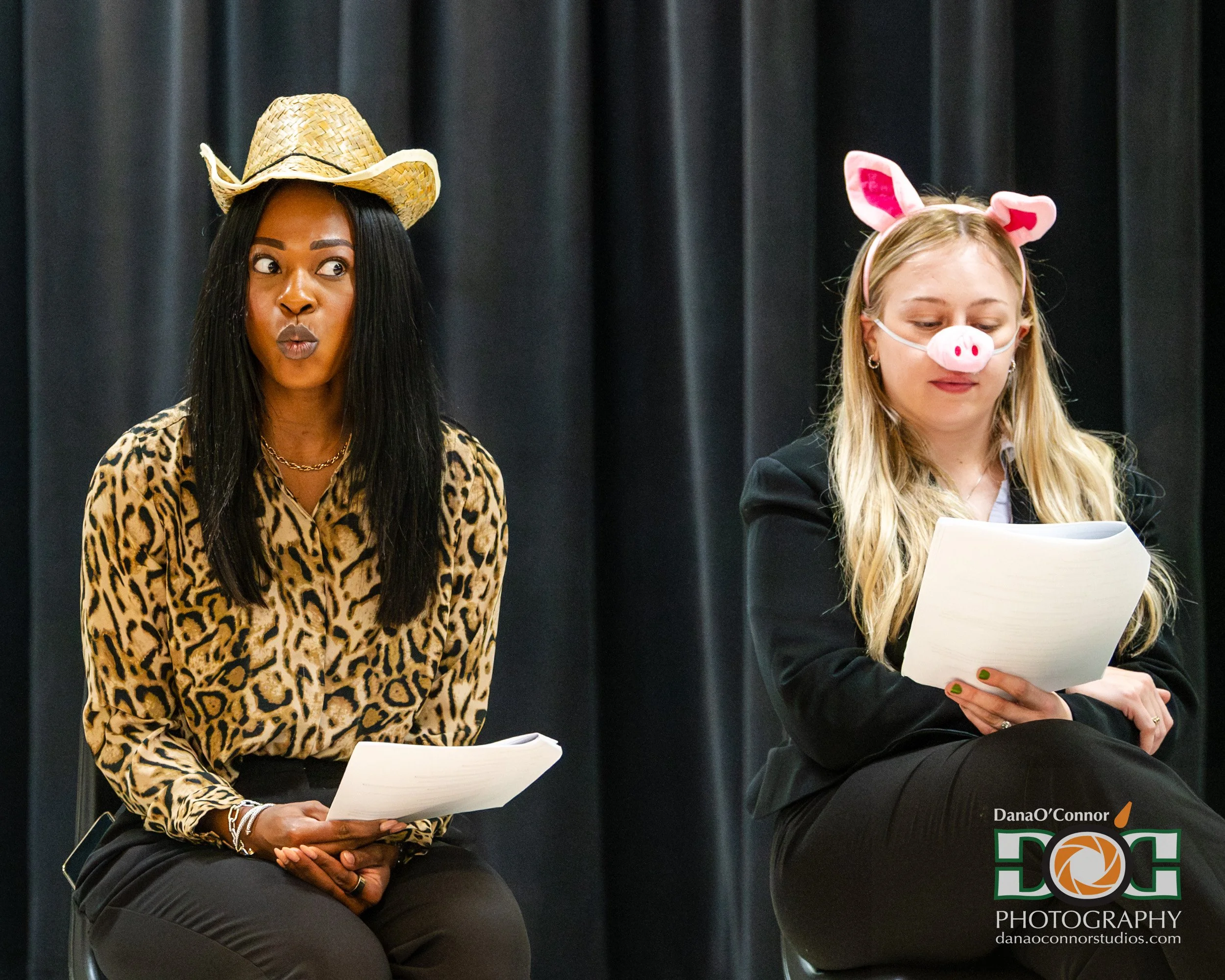 Two women sitting on stage with black curtains behind them. The woman on the left is wearing a leopard print blouse, a straw cowboy hat, and jewelry. The woman on the right is wearing a pig nose and ears headband, reading from a paper. Both are seate