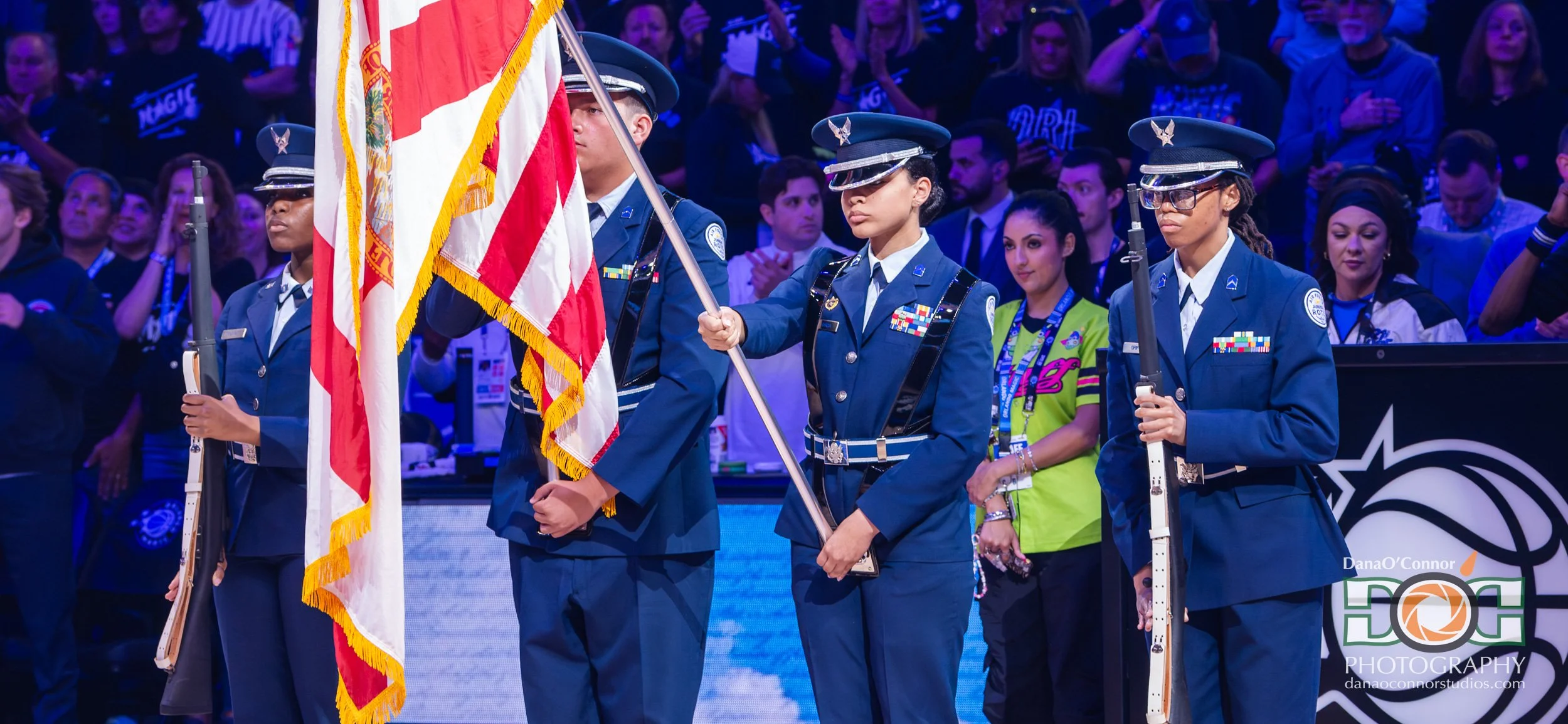 Four individuals in military uniforms holding rifles and a flag at a ceremony, with an audience in the background.