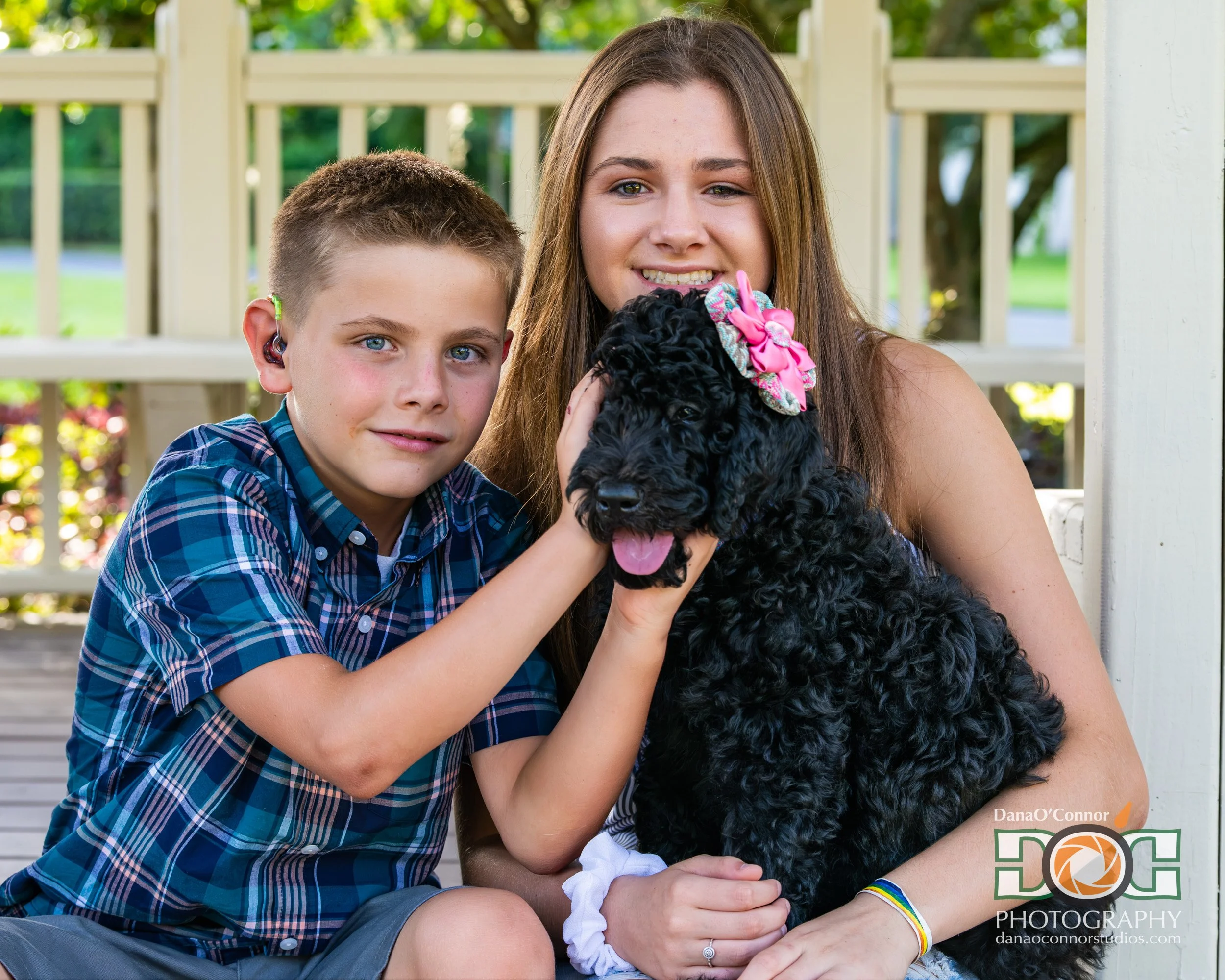 Two kids and a dog sitting outdoors, with a white fence and greenery in the background. The boy has short light brown hair and blue eyes, wearing a blue plaid shirt. The girl has long brown hair and green eyes, wearing a black curly-haired dog with a