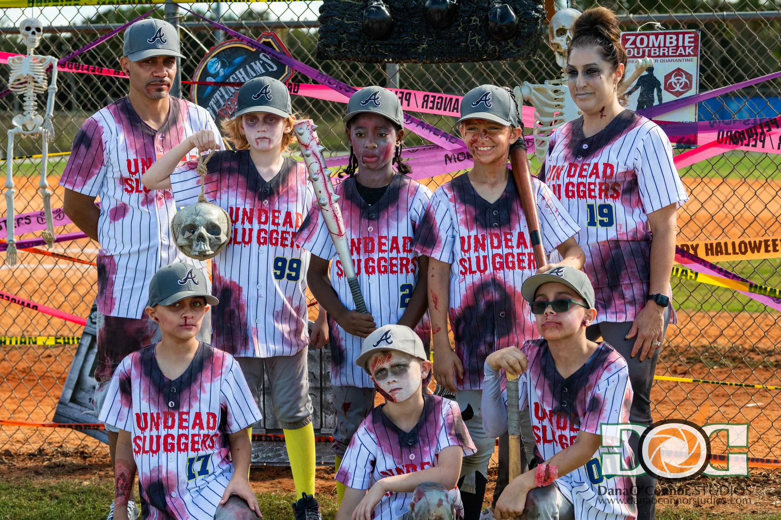 Apopka Little League's 9U Royals dressed as zombies with blood and dirt on their faces and uniforms, standing behind a chain-link fence decorated with pink caution tape and Halloween decorations, during daytime.