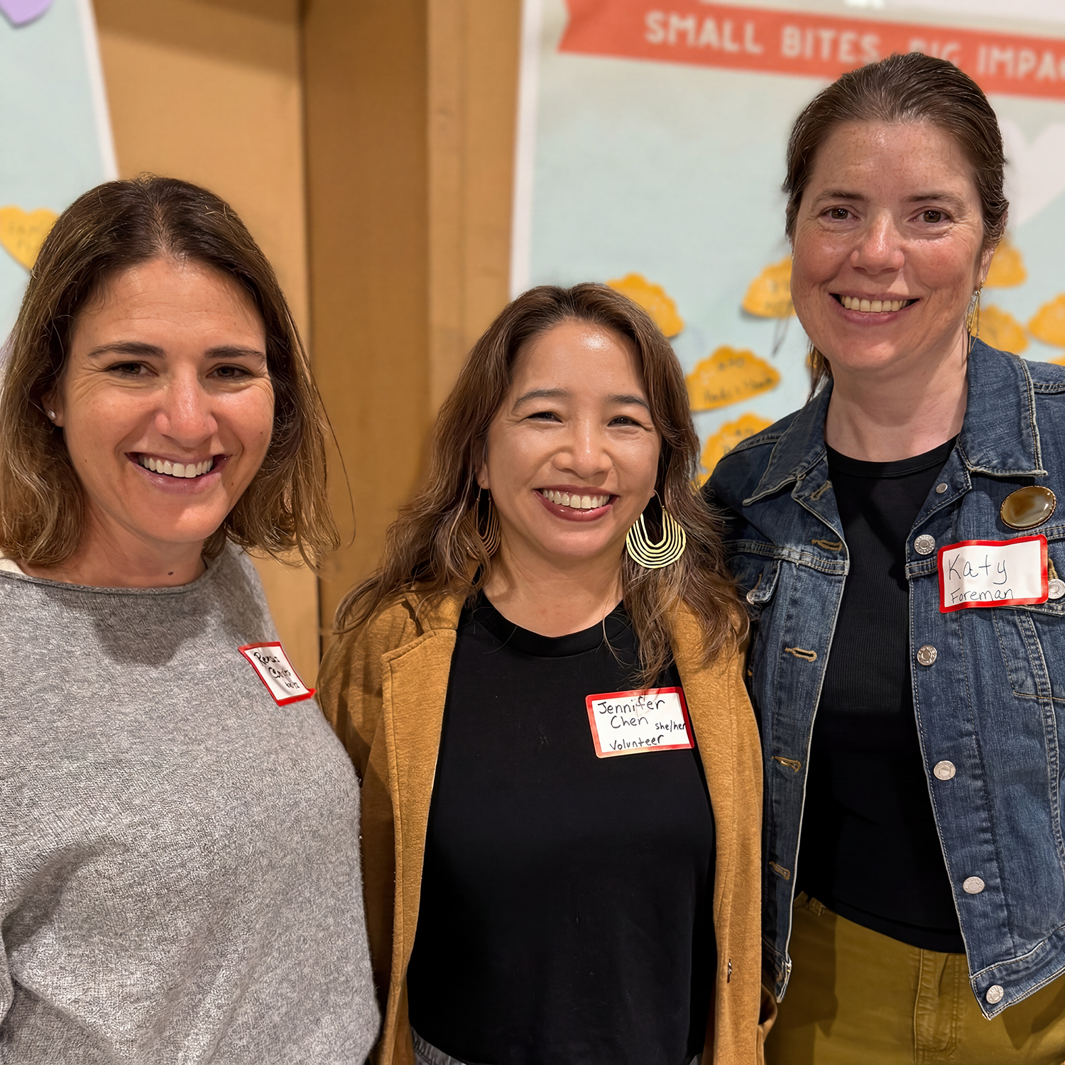 Three people smiling, standing close together at an indoor event: Rebecca Bauer-Kahan, Jennifer Chen, and another woman. They wear name tags and casual clothing. The background has a colorful poster with text.