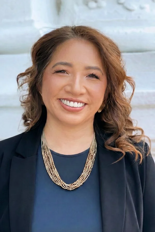 Jennifer Chen, candidate for re-election: an Asian American woman with shoulder-length curly hair, smiling, wearing a navy blazer, navy top, and layered beaded necklaces, standing against a light-colored outdoor background
