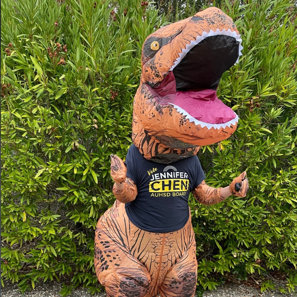 Person in a dinosaur costume with a Jennifer Chen campaign t-shirt standing outdoors in front of green bushes.
