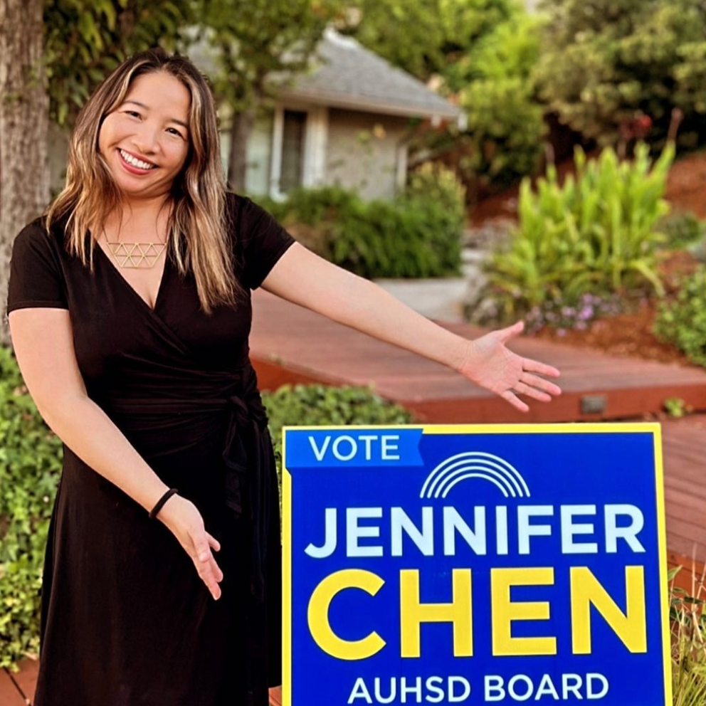 Woman smiling and pointing at a campaign sign for Jennifer Chen, running for AUHSD Board, outdoors in a garden setting.