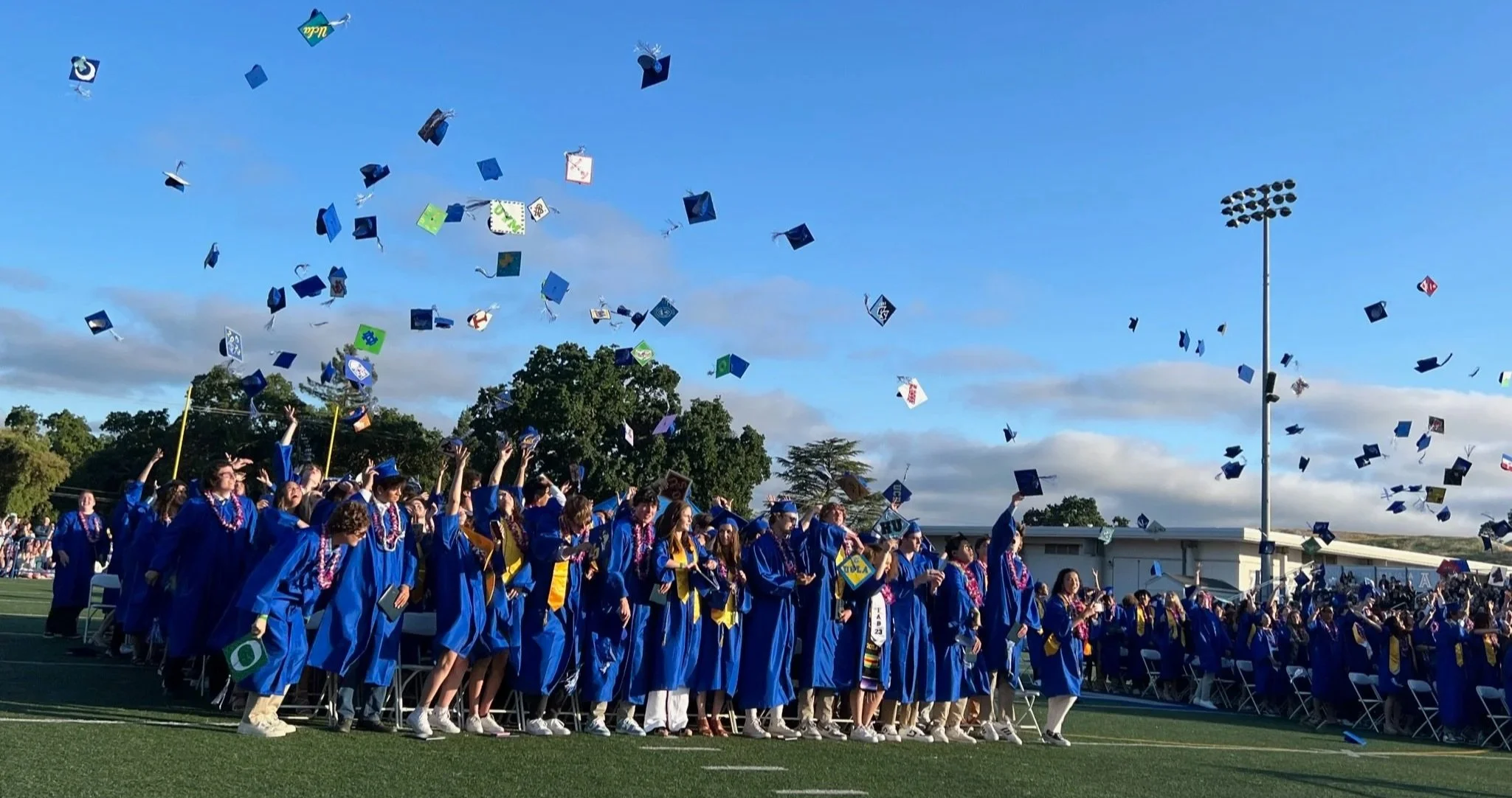 High school graduates in blue caps and gowns celebrate outdoors, tossing their caps into the air during graduation ceremony on a football field under a clear blue sky.