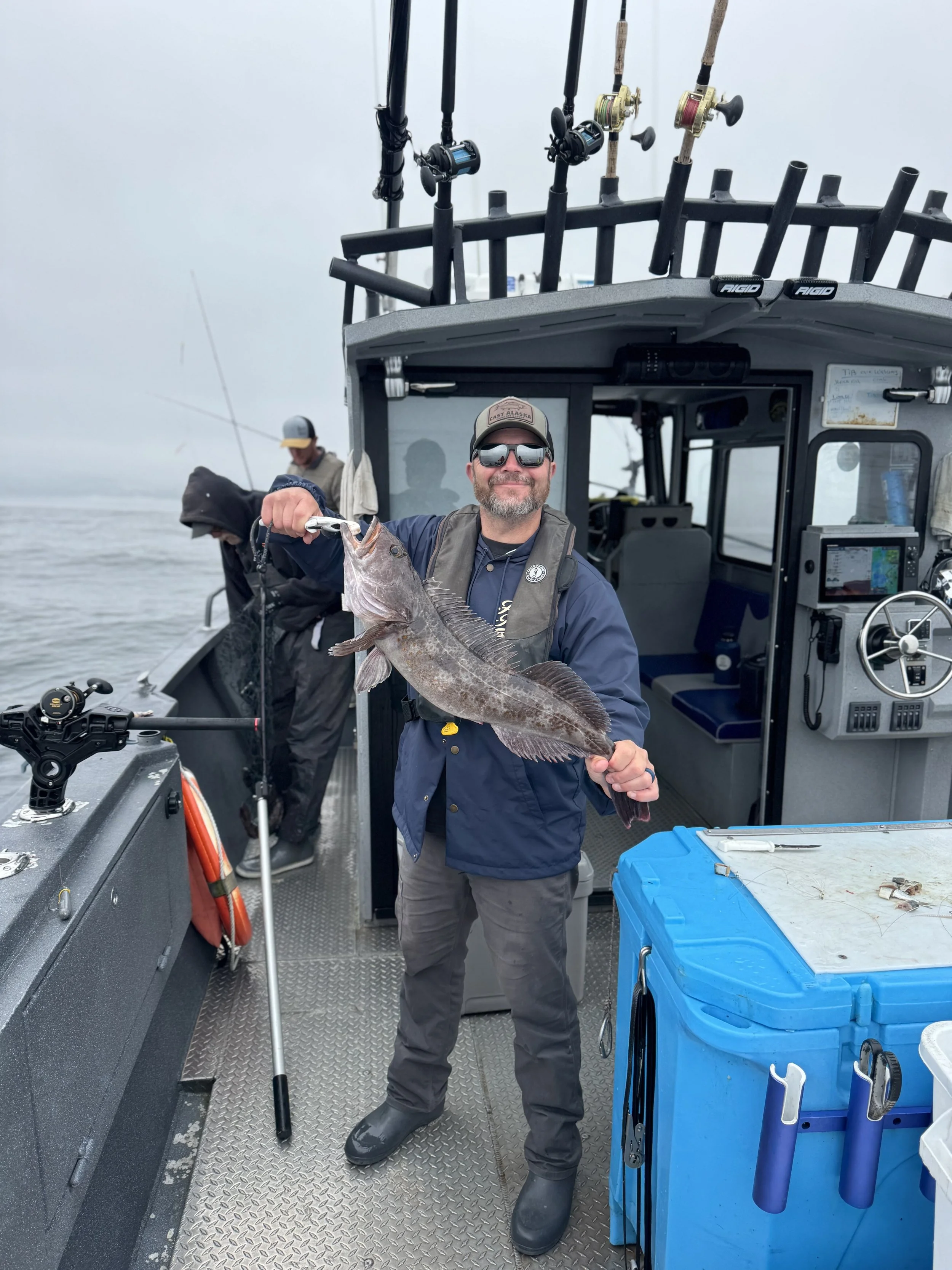 A man holding a large fish on a boat with fishing gear and other people in the background, on a cloudy day.