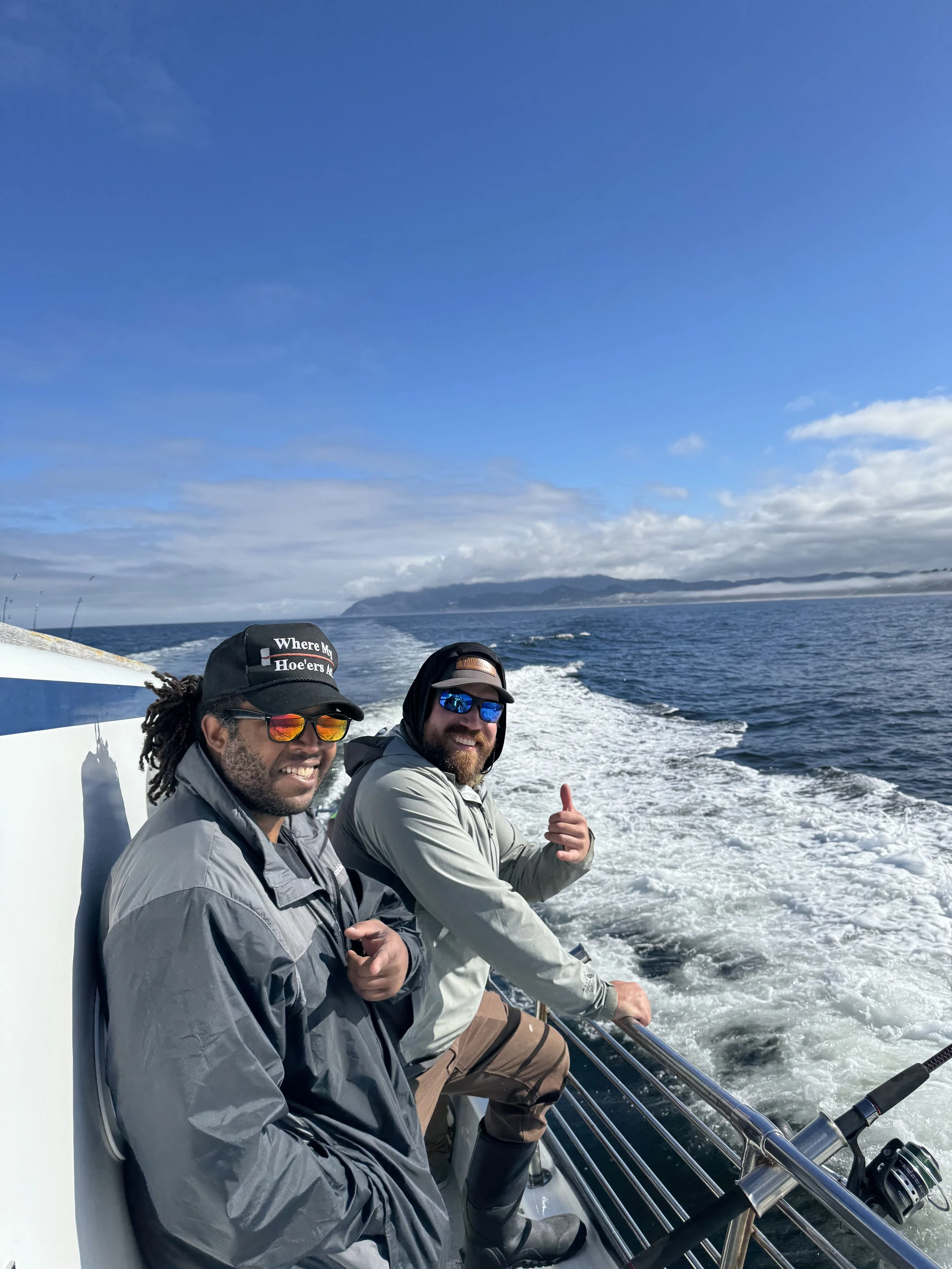 Two men on a boat smiling, one giving a thumbs up, in a large body of water with land in the distance and a blue sky