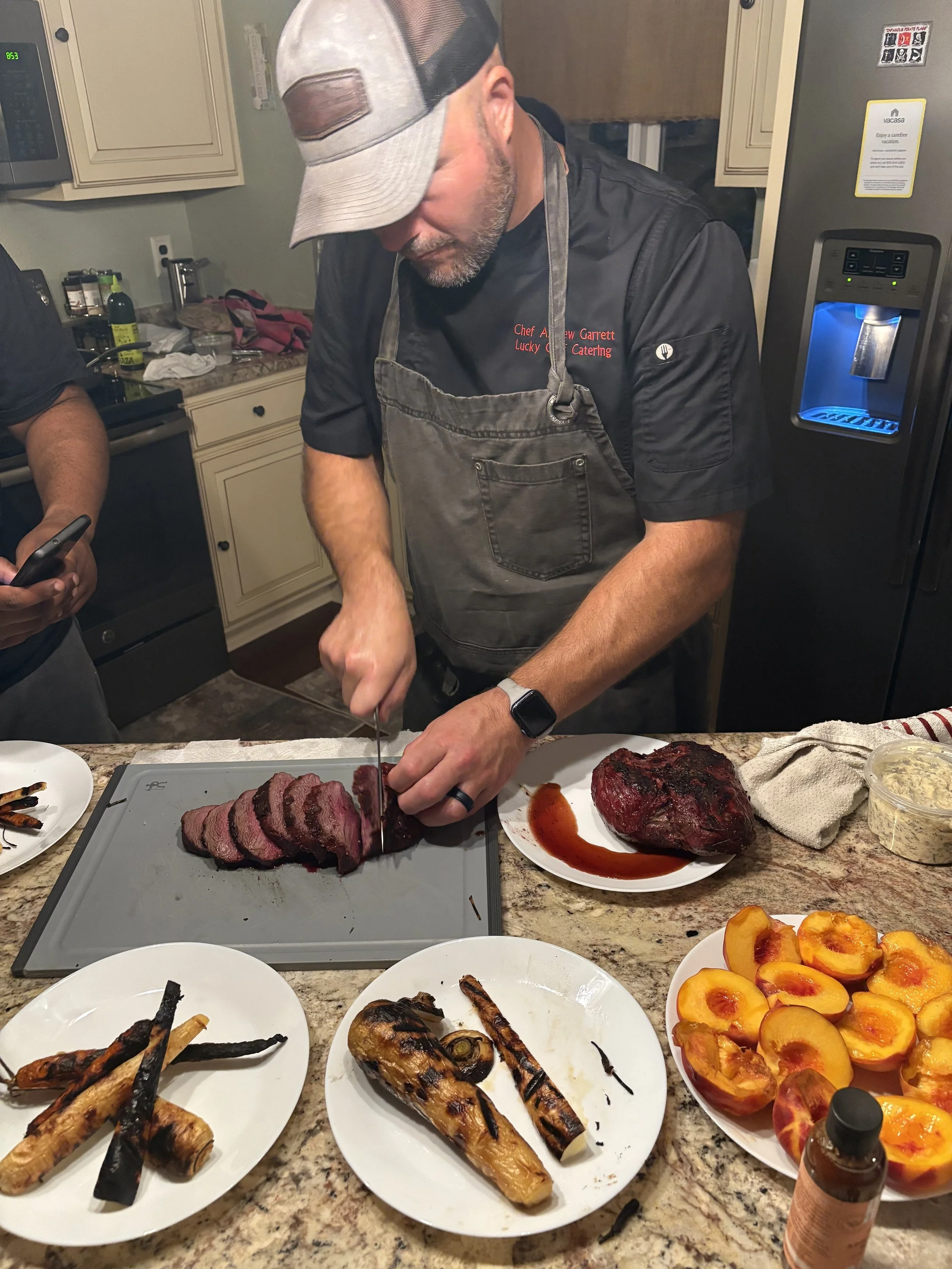 Man in a black chef's coat and gray apron is slicing a cooked steak on a cutting board in a kitchen, surrounded by plates of grilled vegetables, peaches, and a large cooked piece of meat