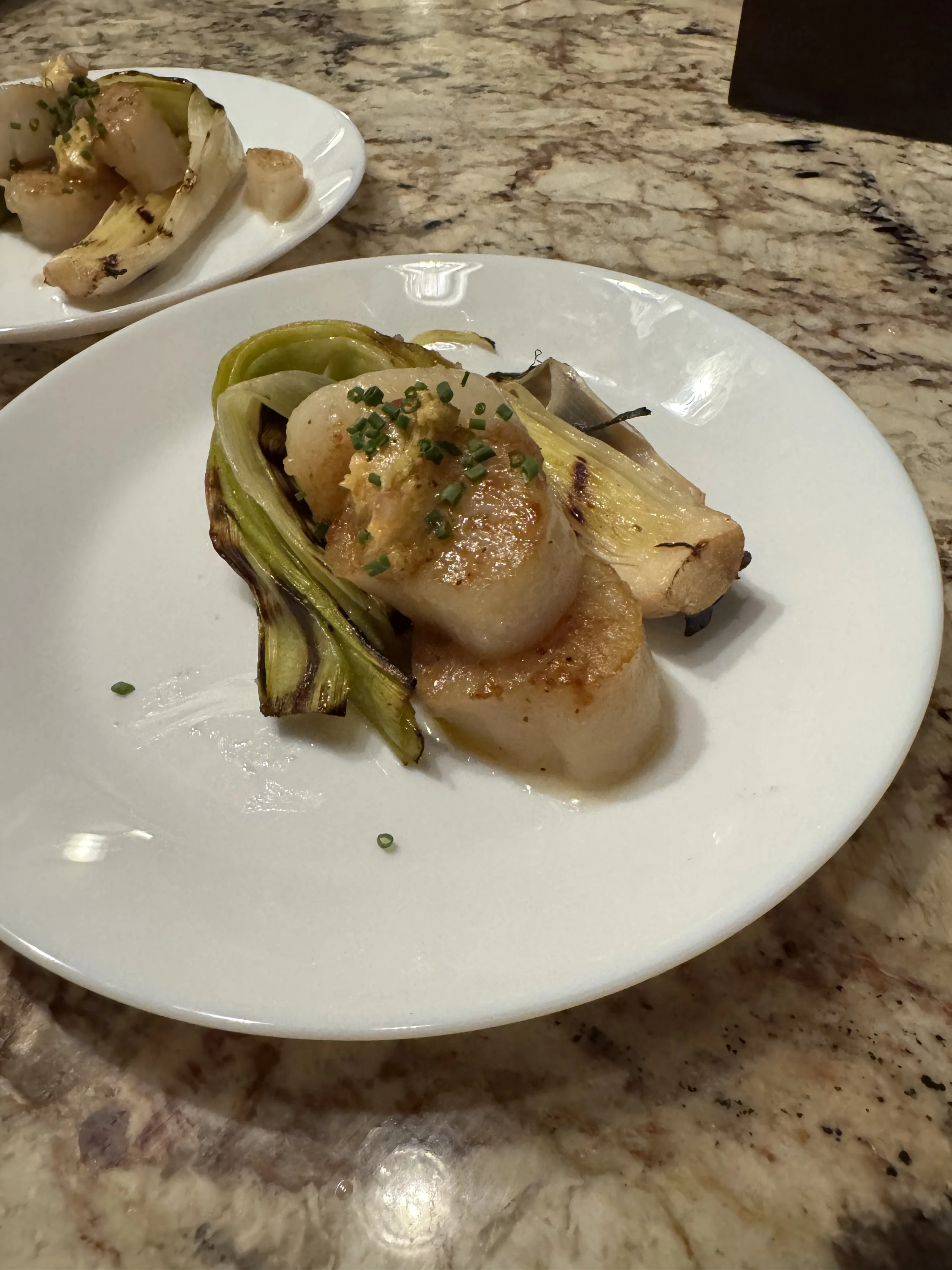 A plate of grilled scallops with green vegetables, garnished with chopped chives, on a white plate, with a bowl of more grilled vegetables in the background, placed on a marble countertop.