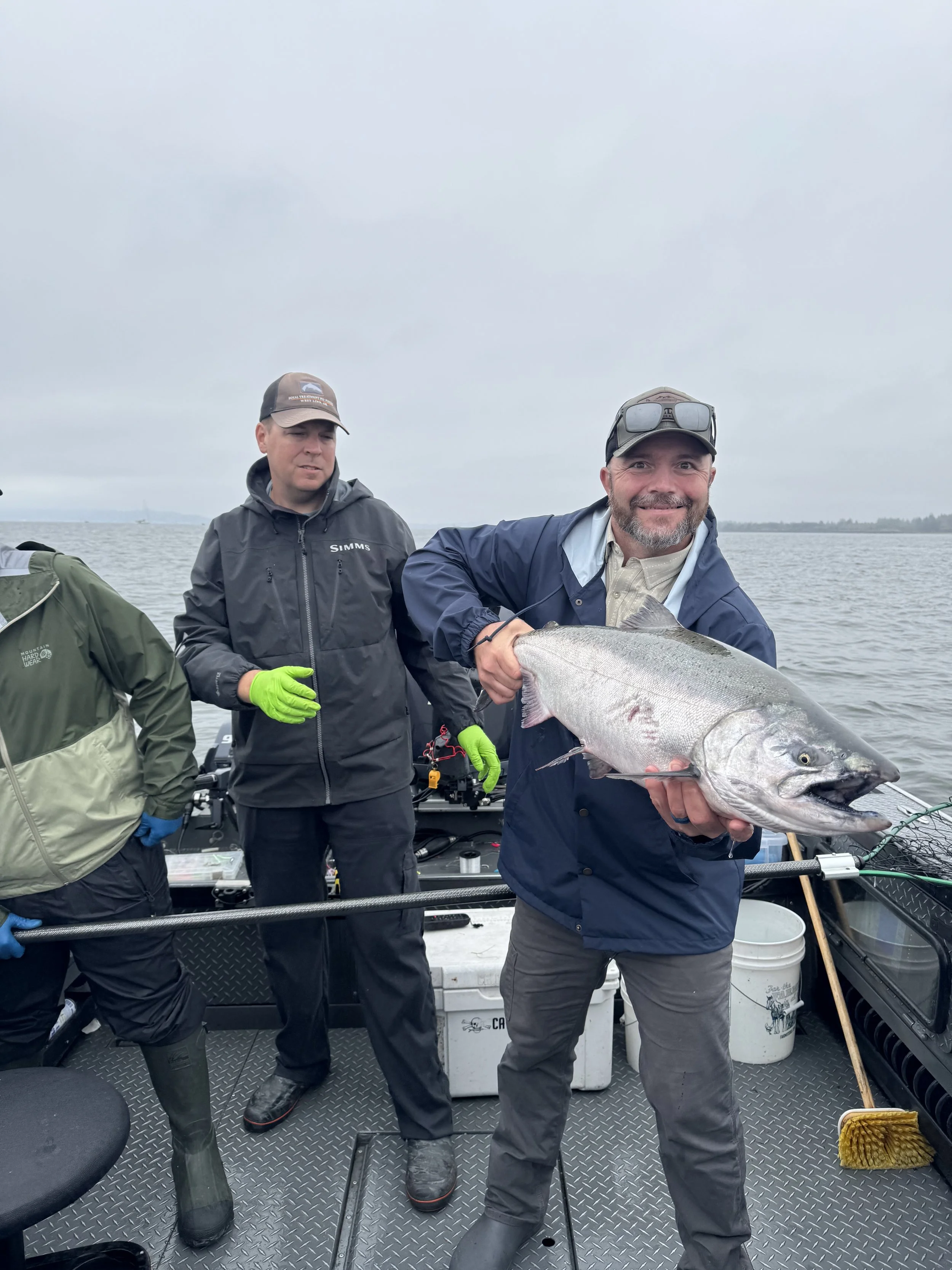 Man holding a large fish on a boat with two other men, all wearing outdoor gear, in a cloudy outdoor setting on the water.
