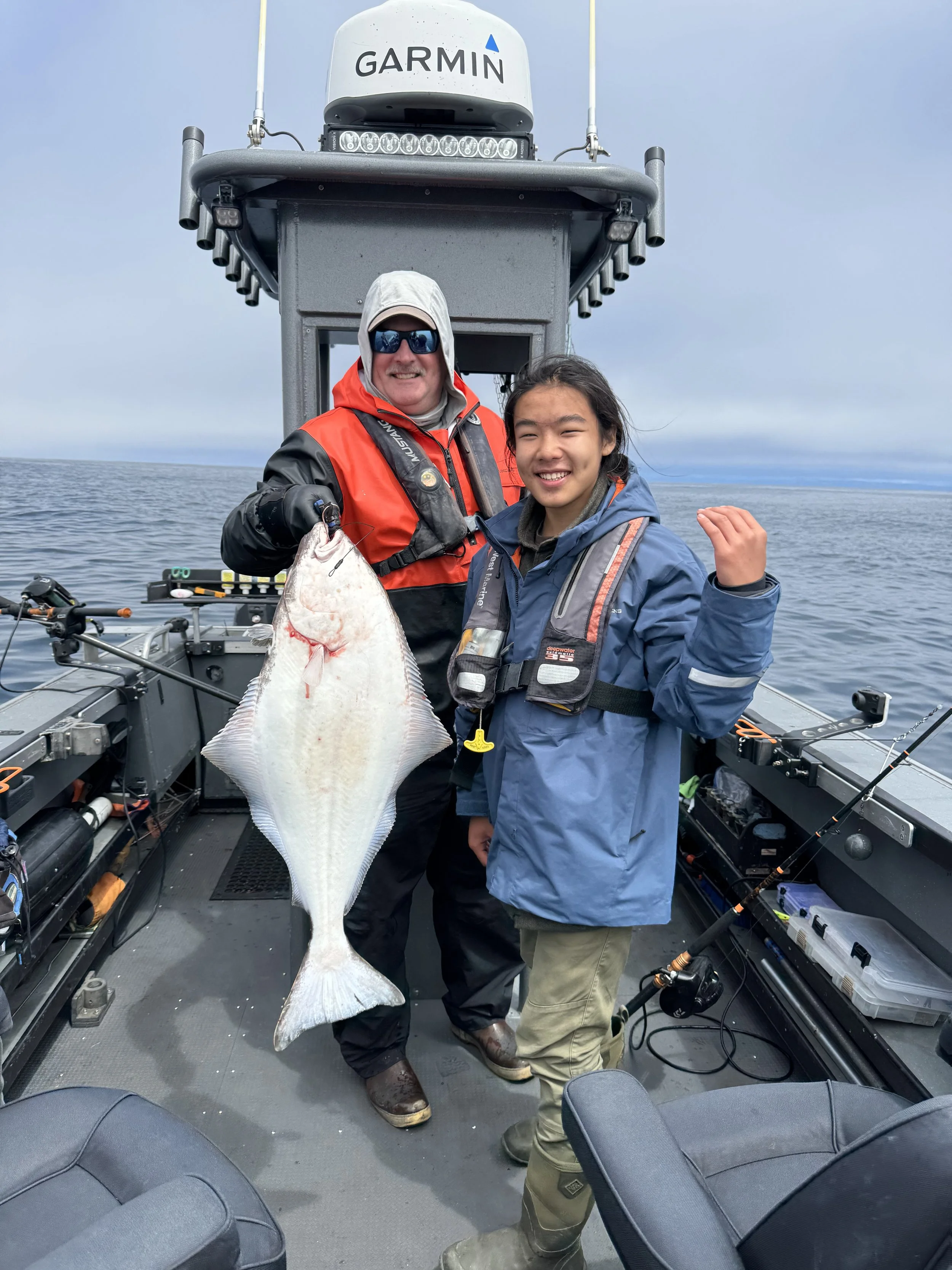 Two people on a boat, one holding a large fish, with a gray ocean and cloudy sky in the background.