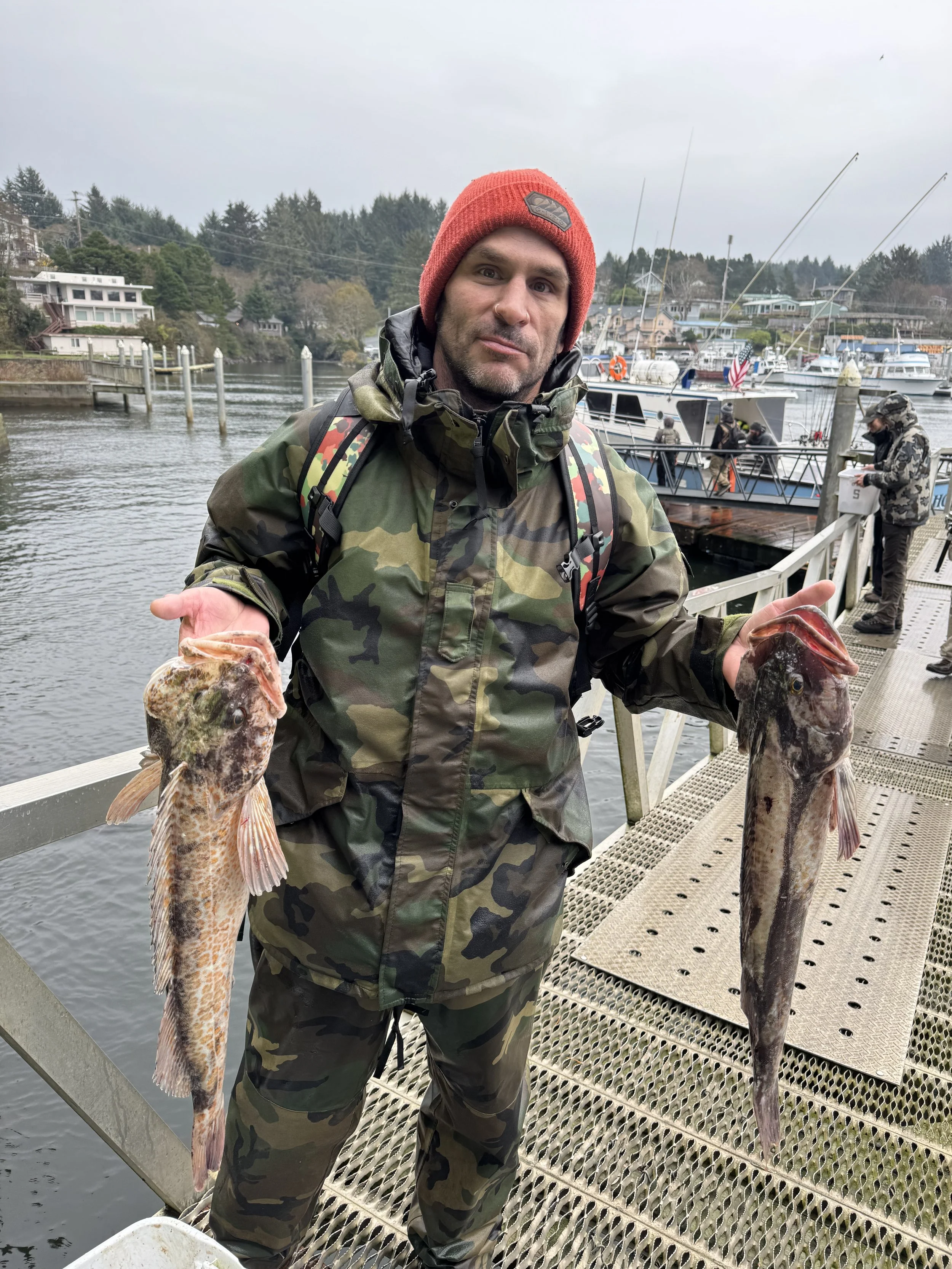 Man standing on a dock holding two large fish, wearing camo clothing and a red beanie, with boats and other people fishing in the background at a marina.