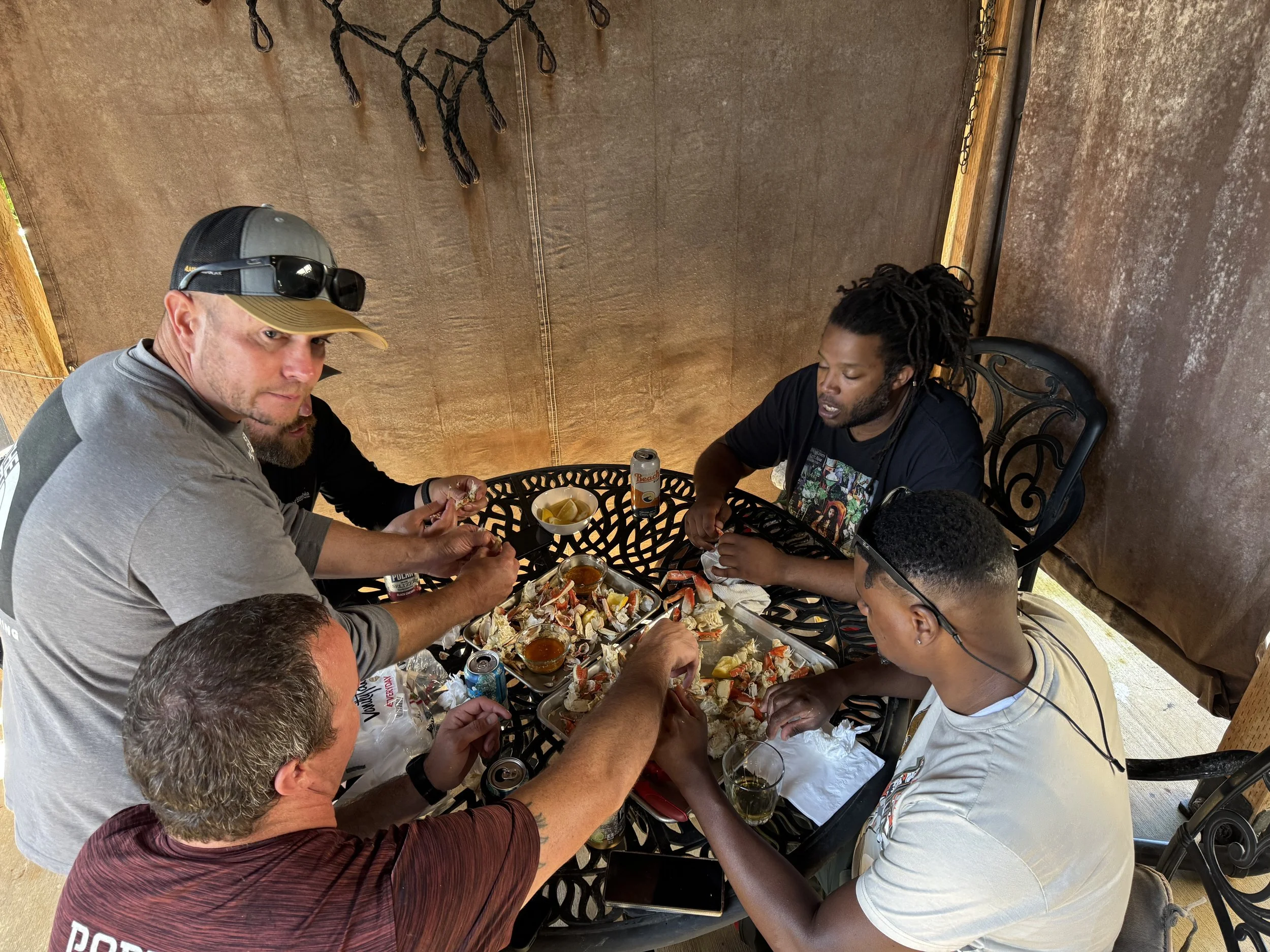A group of five people sitting around a metal outdoor table, sharing a seafood feast with crab legs, drinks, and dipping sauces under a shaded canopy.