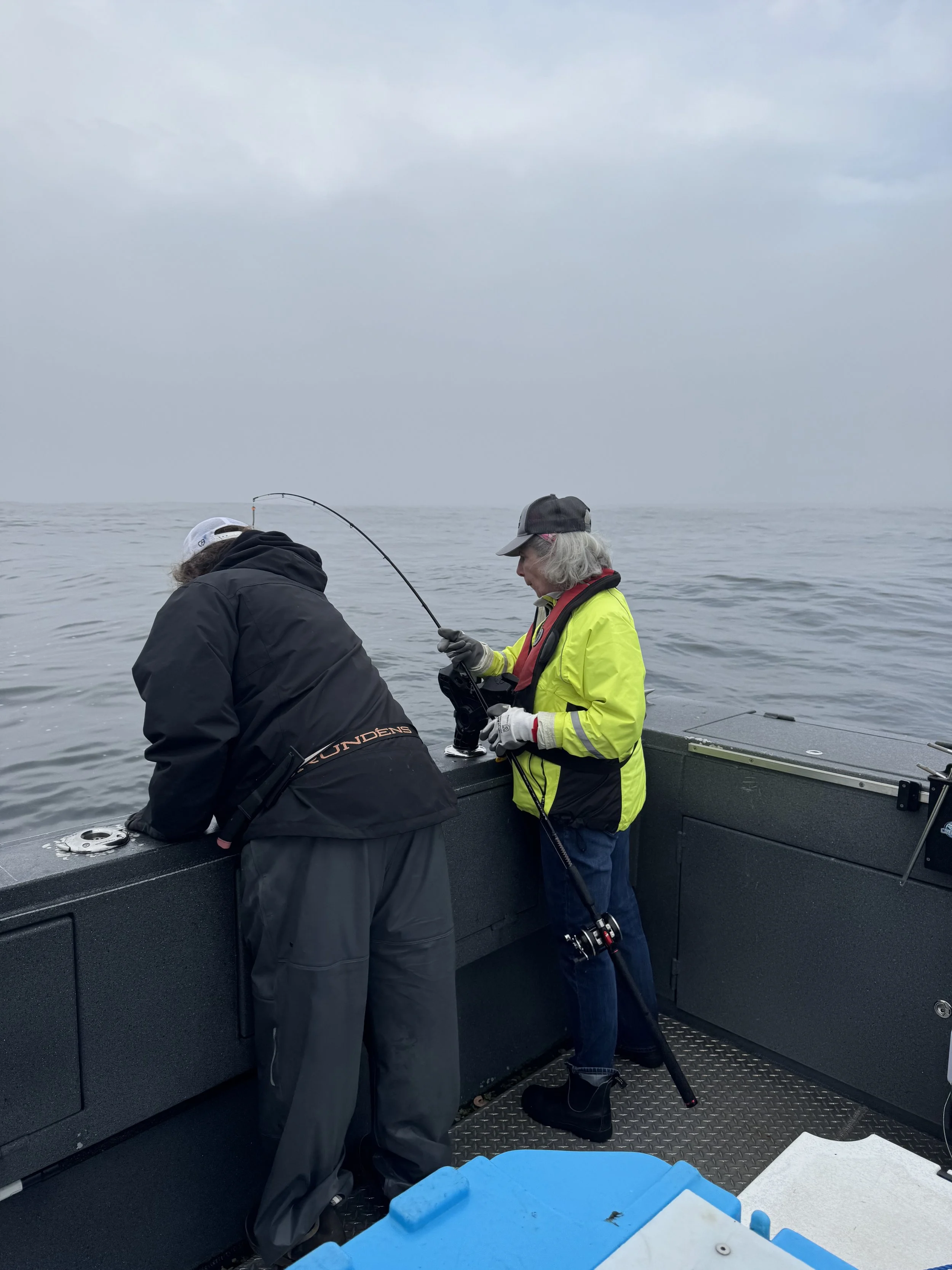 Two women on a boat fishing in a cloudy, open water setting.