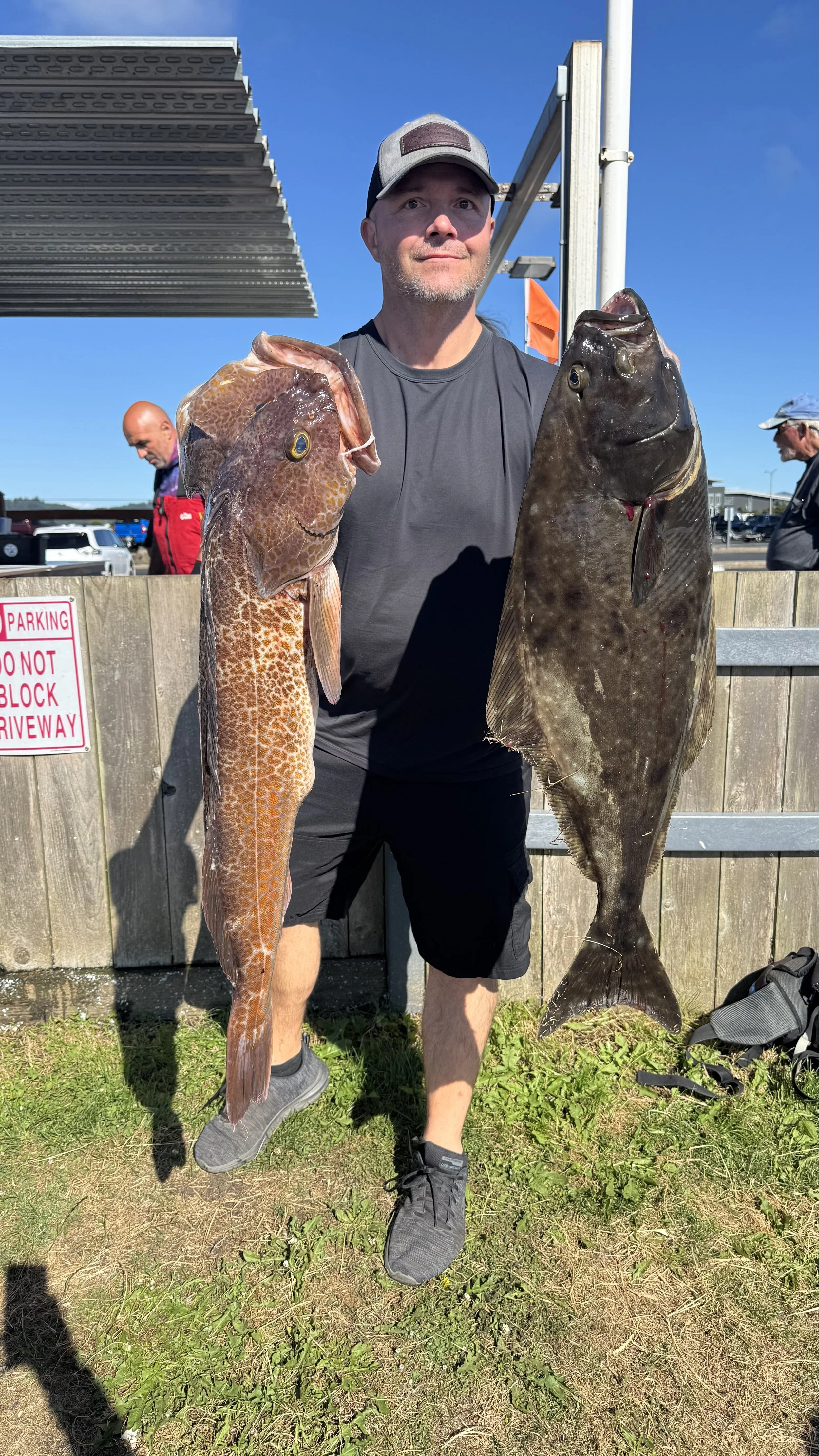 Man holding two large fish, one in each hand, outdoors at a fishing site with a blue sky and people in the background.