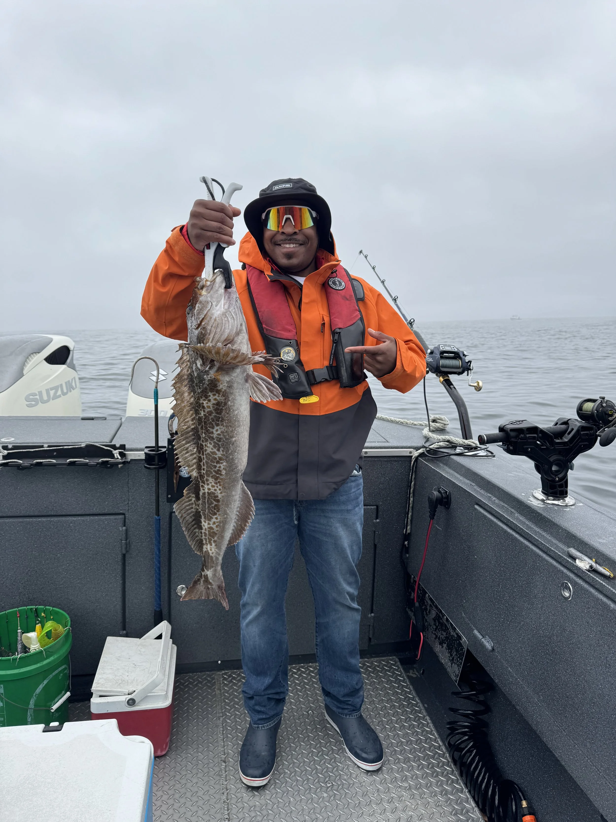 Man on a boat holding a large fish he caught, wearing an orange jacket and sunglasses, with fishing gear and the ocean in the background.