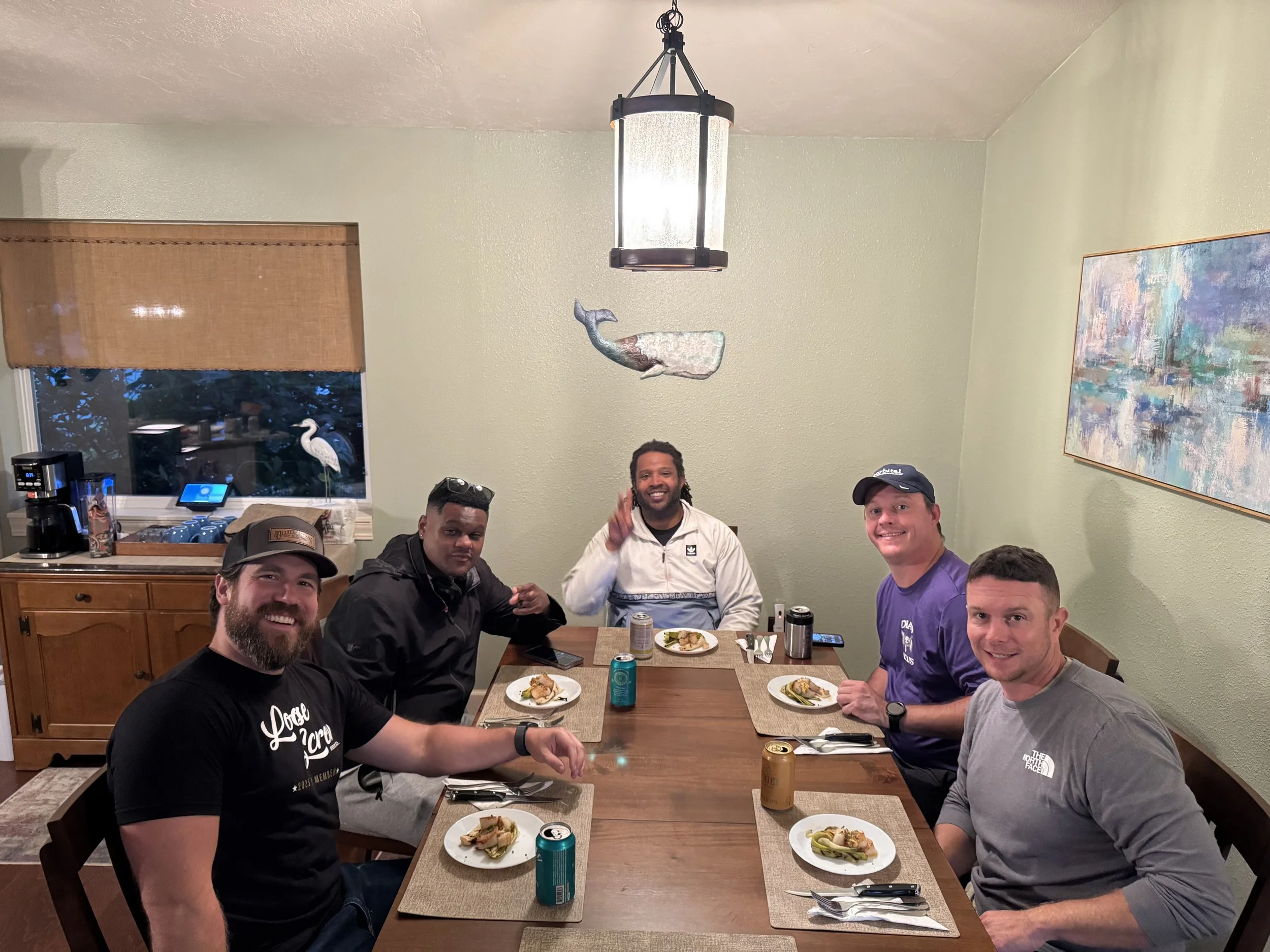 Five men sitting around a dining table with plates of food, cans of soda, and utensils, smiling and posing for a photo in a dining room with green walls, a large abstract painting, a whale decoration hanging from the ceiling, and a window showing a b