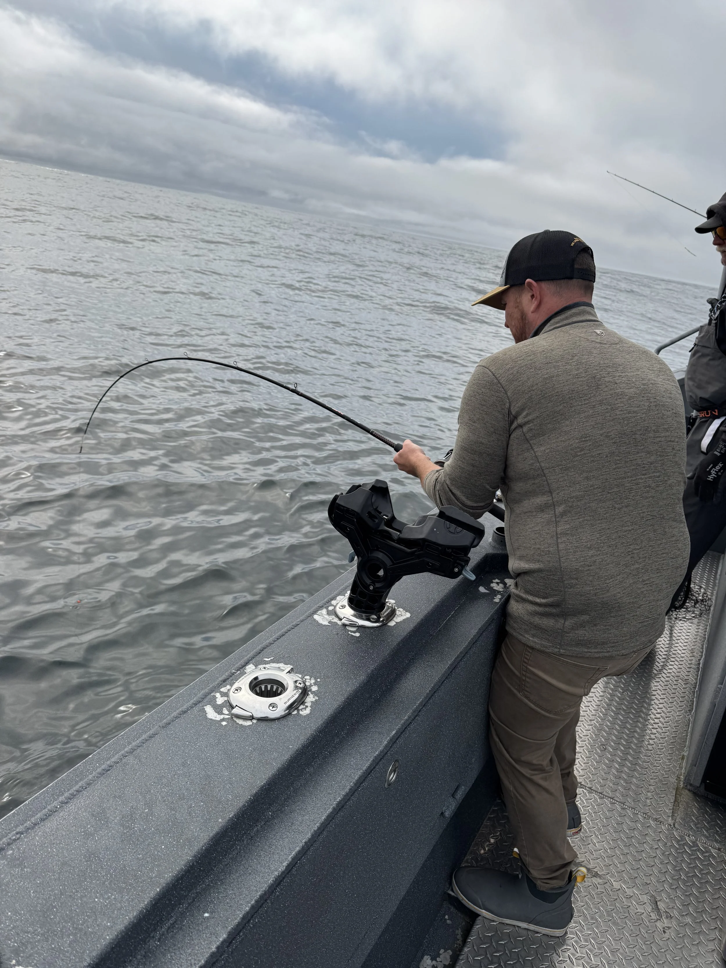 A man fishing from a boat on a cloudy day with another person standing beside him, with water and cloudy sky in the background.