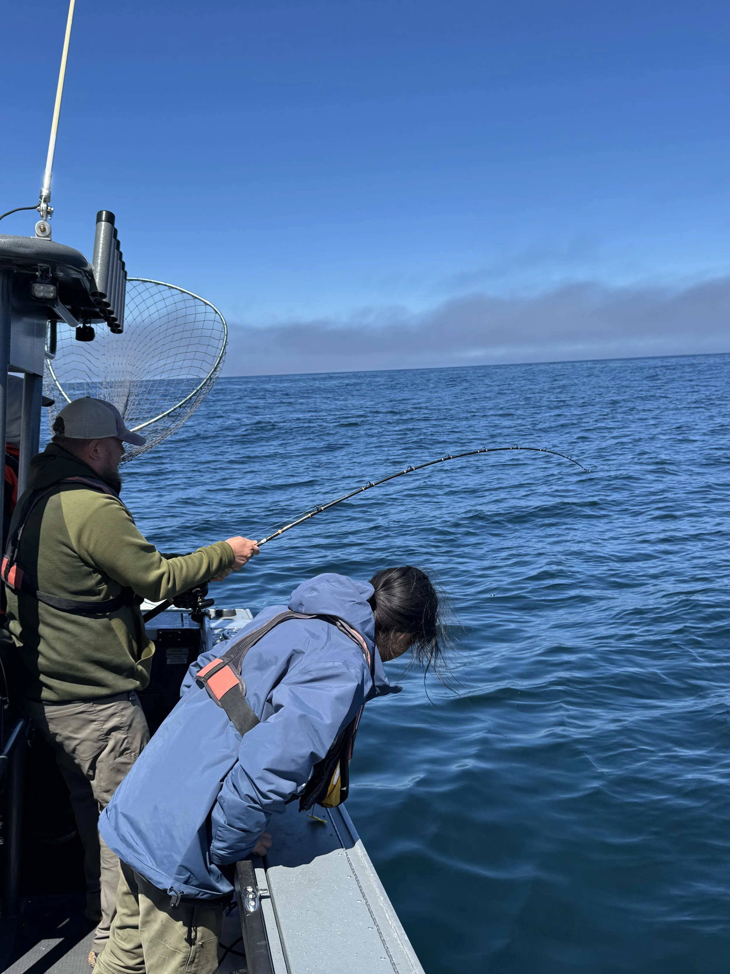 Two people on a boat fishing in the ocean under a partly cloudy sky.
