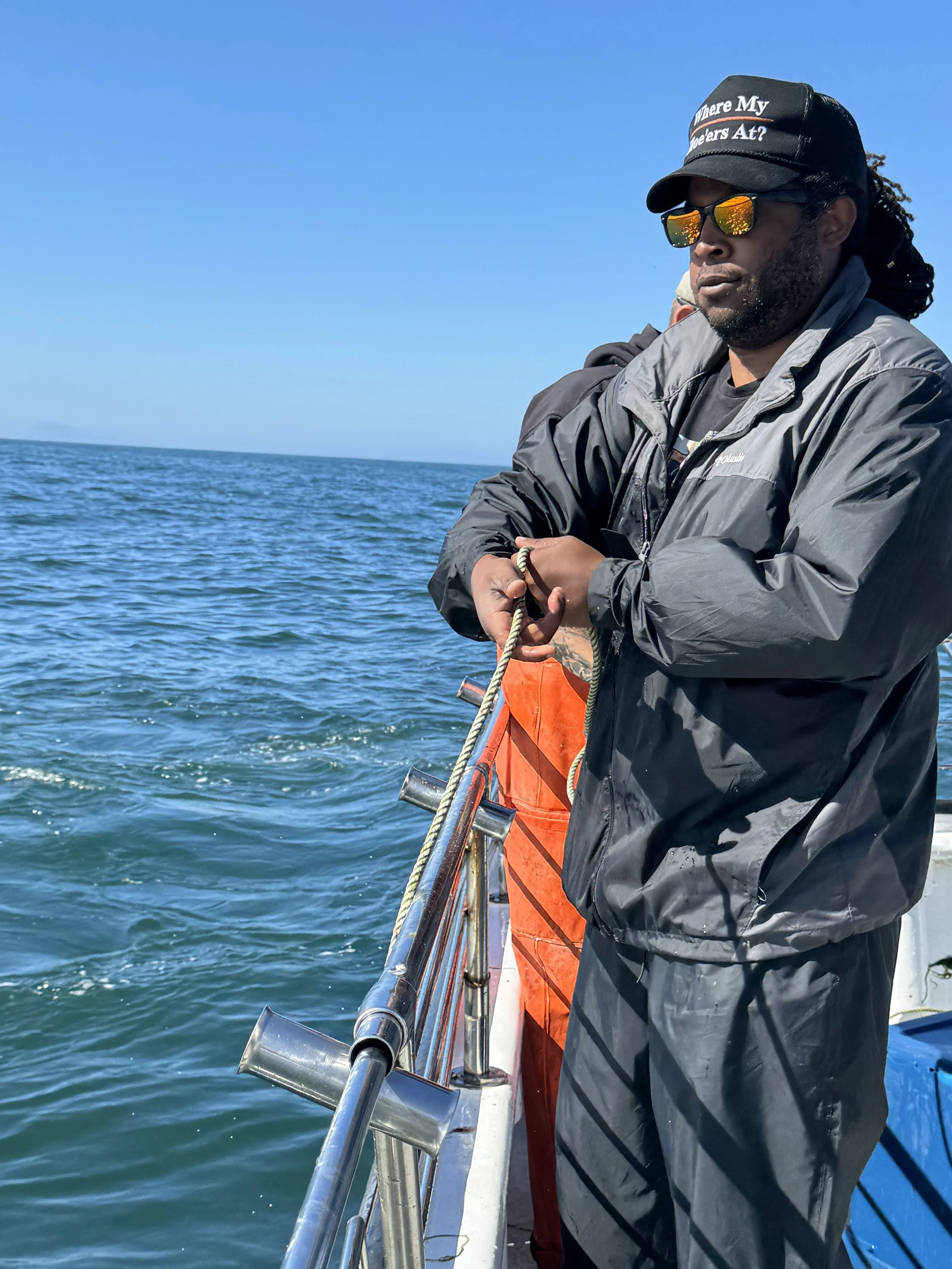 A man standing on a boat, fishing in the ocean, wearing a black cap with text and sunglasses, dressed in a gray jacket and black pants.