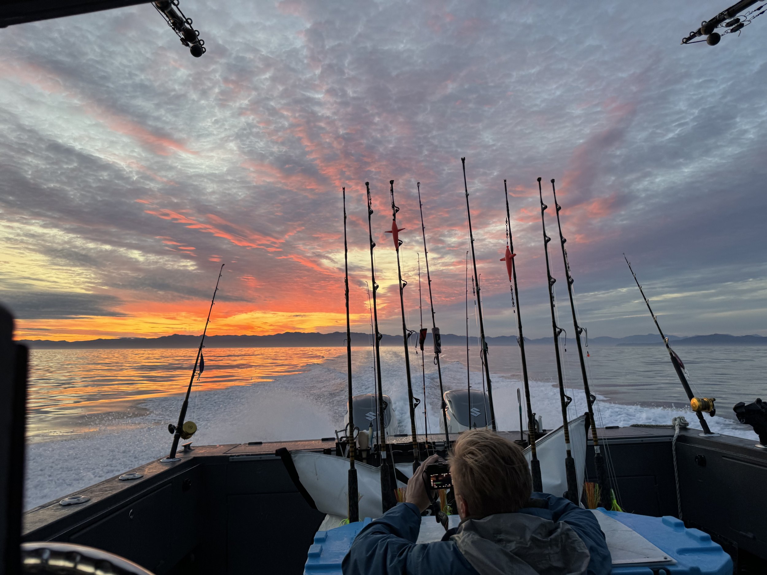 Person on a boat taking a photo of a colorful sunset sky with fishing rods at the stern, over calm water with distant mountains.