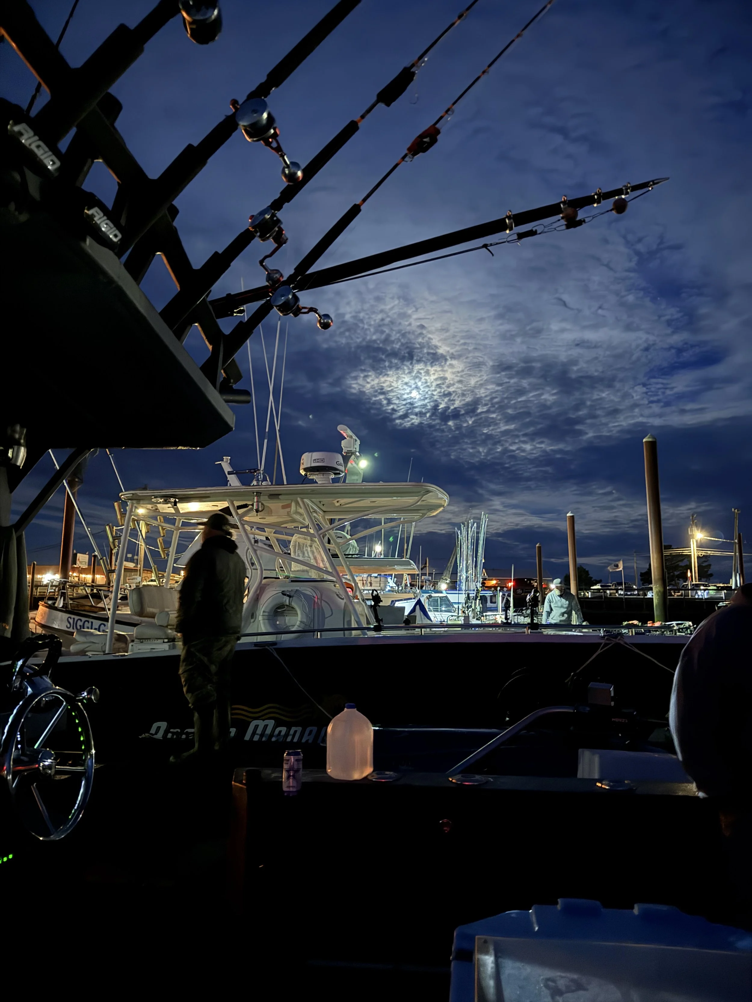 Nighttime scene at a marina with boats docked, fishing rods mounted on a boat, and a dark sky with clouds and the moon.
