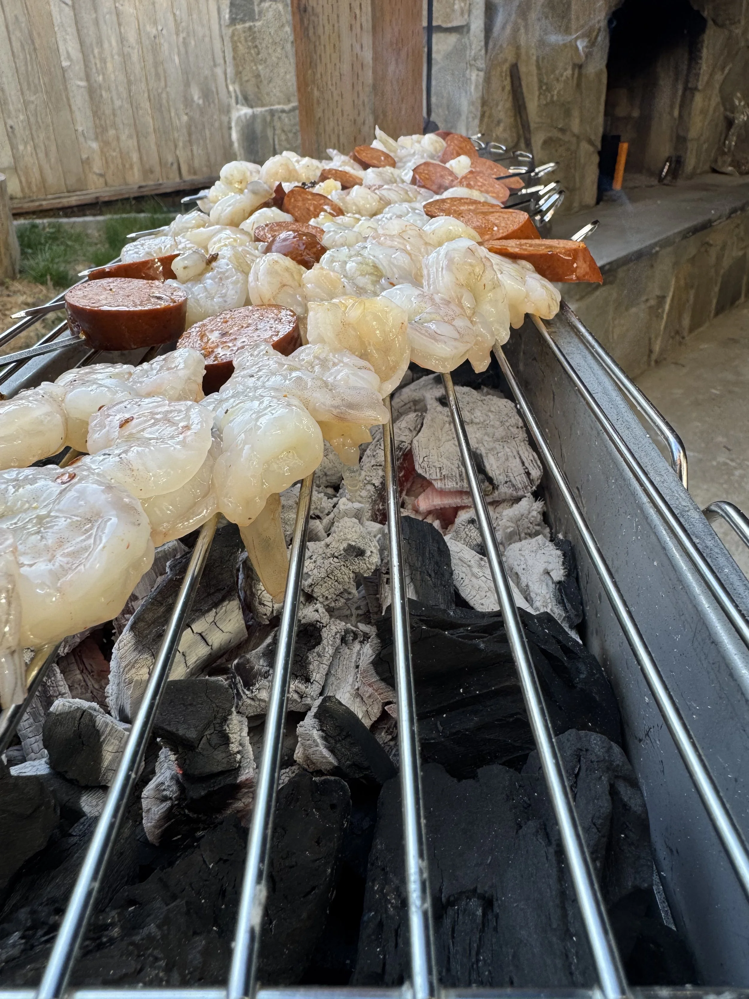 Skewers with raw shrimp and sausage cooking over charcoal in an outdoor grilling area.
