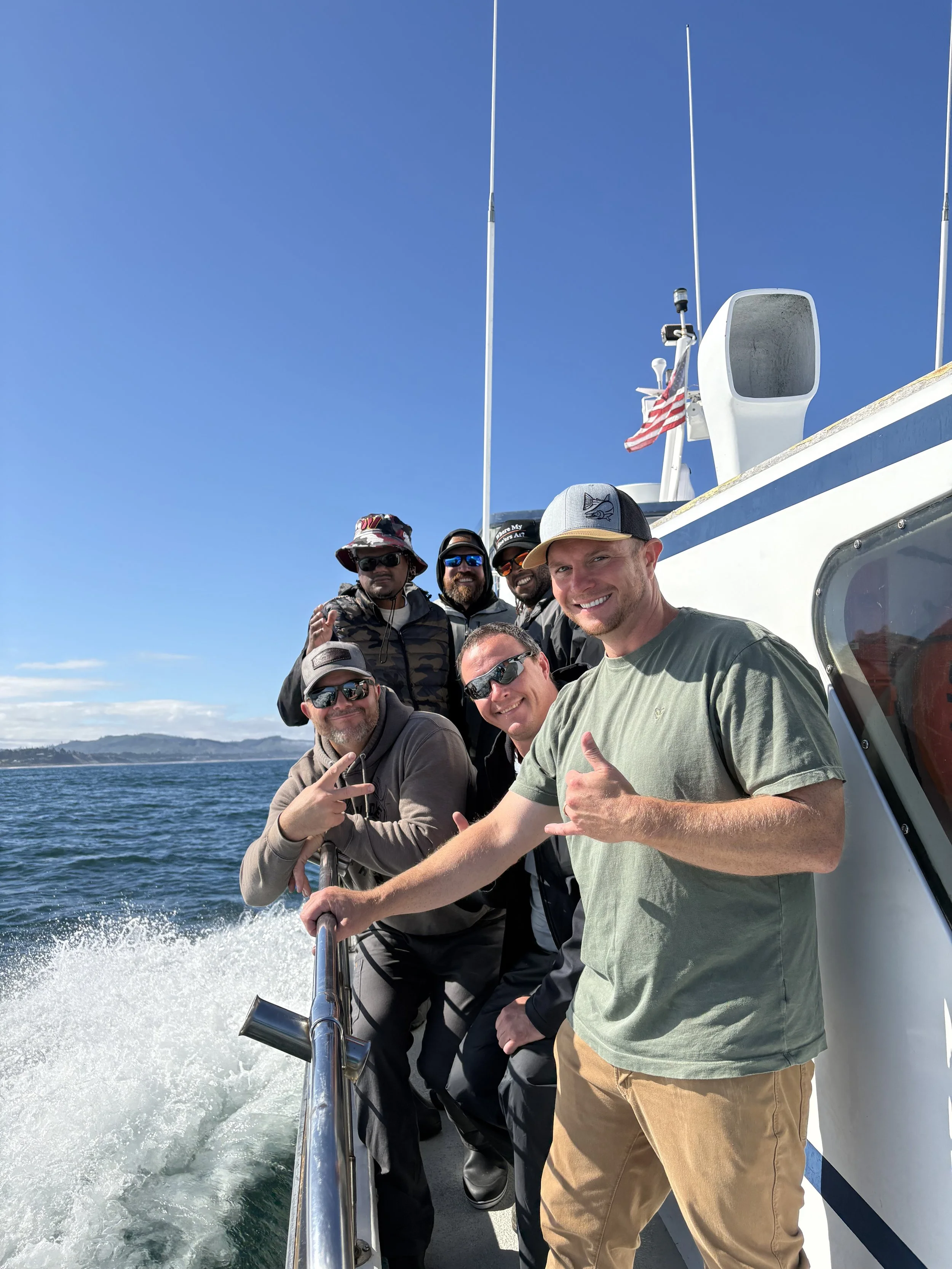 Group of six men on a boat, smiling and making hand gestures, with a blue sky and ocean in the background.
