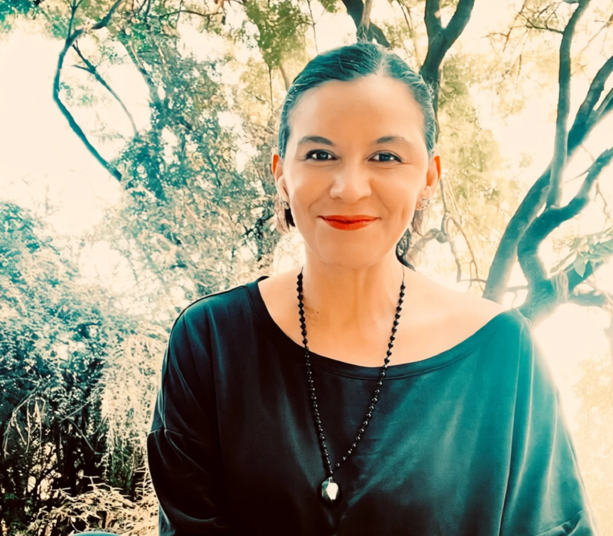 A woman with short dark hair, dressed in a black top with a necklace, smiling outdoors with trees and sunlight in the background.
