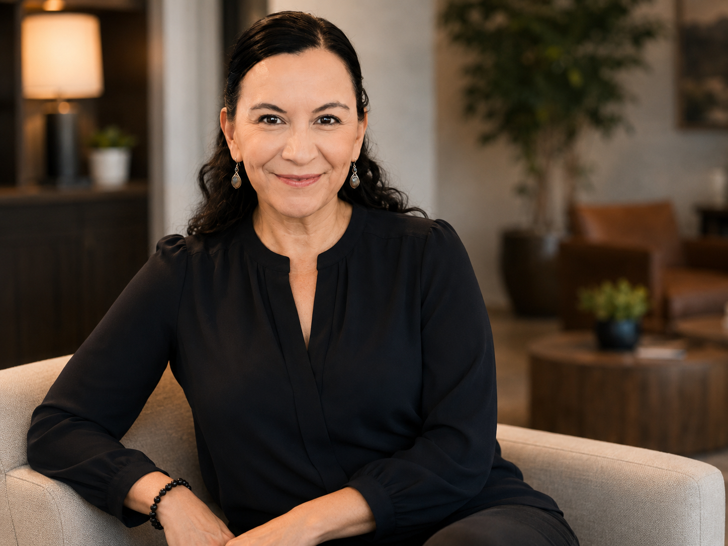 A woman sitting on a beige sofa in a well-lit room, wearing a black blouse and earrings, with a friendly expression.