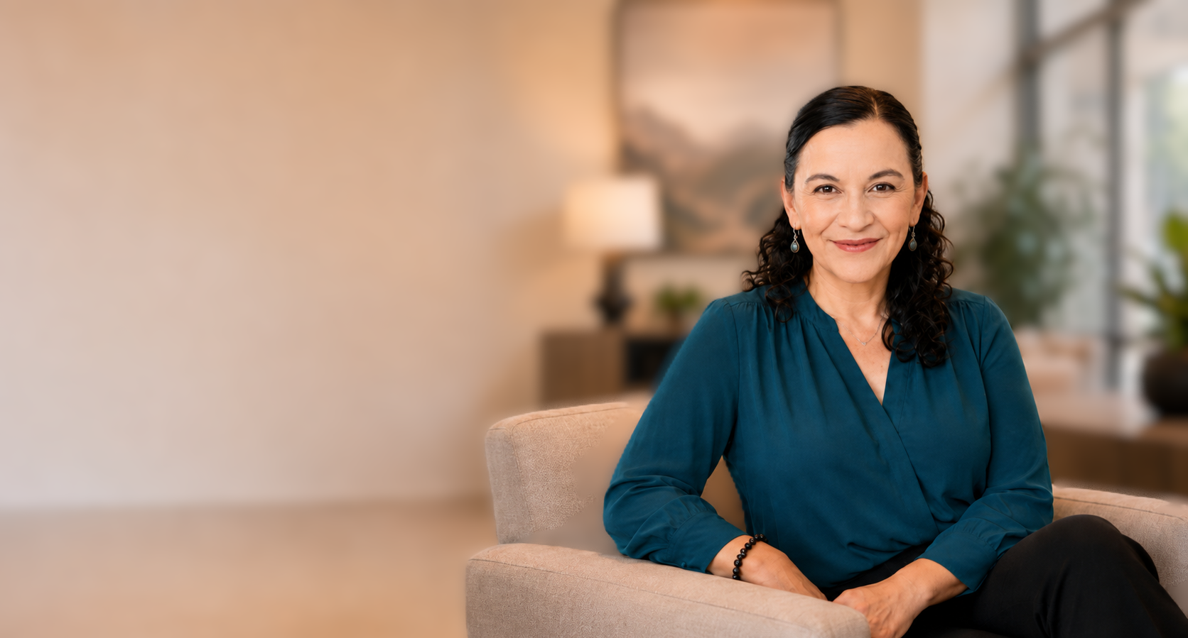 A smiling woman with dark hair and earrings, wearing a teal blouse, sits on a beige couch in a warmly lit room with a lamp, artwork, and plants in the background.