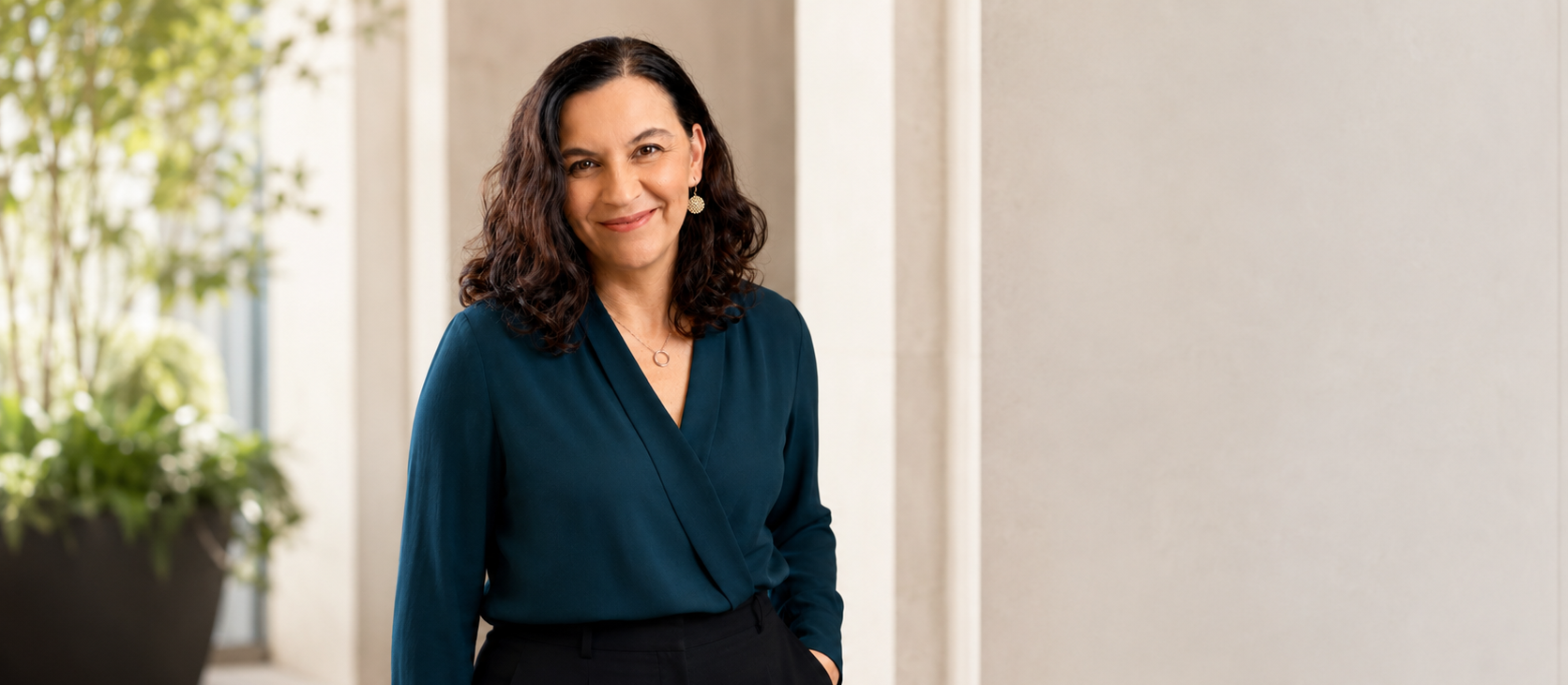 A woman with shoulder-length curly dark hair, wearing a dark teal blouse and black pants, smiling and standing in a well-lit indoor space with a blurred potted plant in the background.