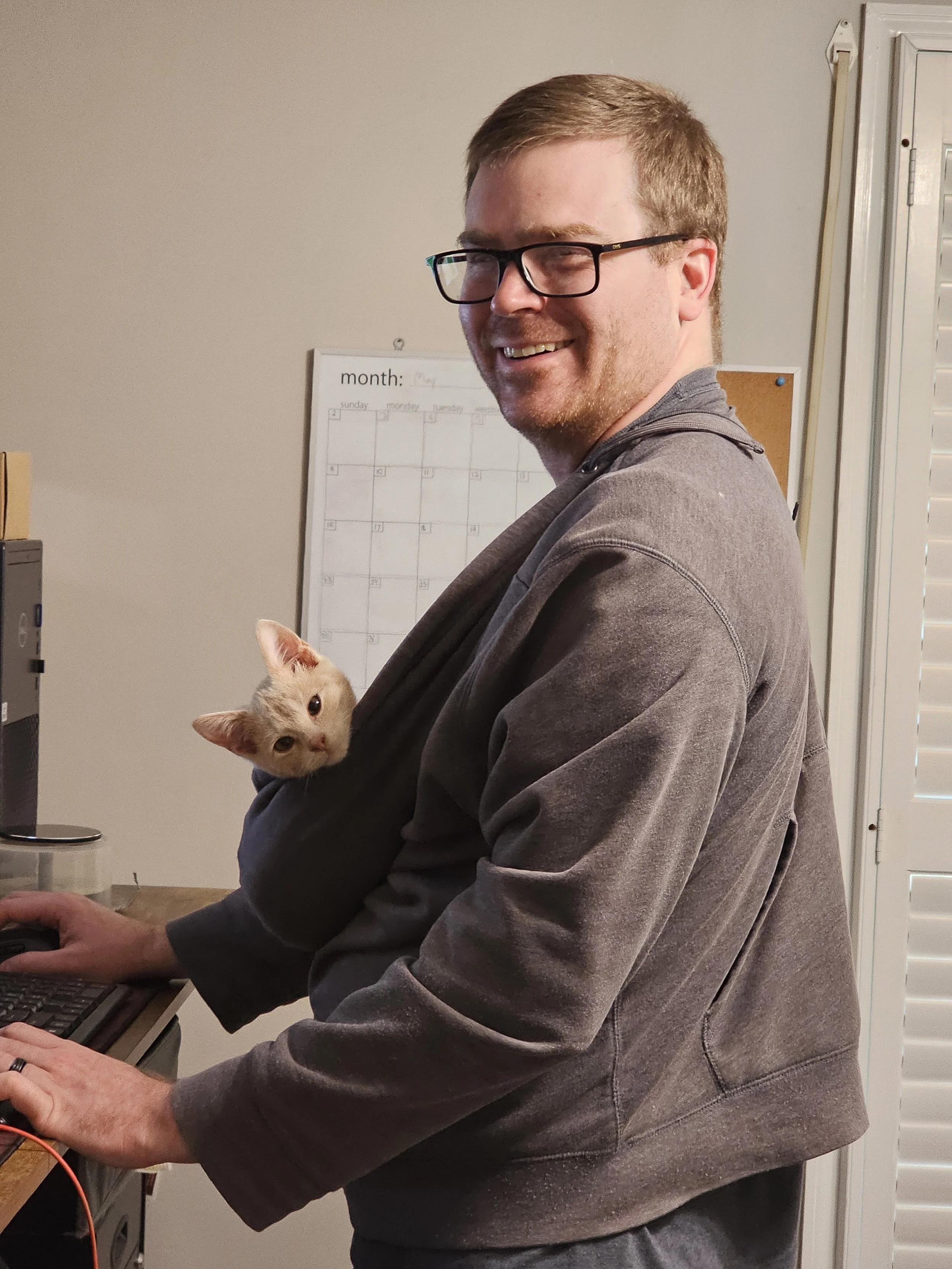 A man with glasses and a gray hoodie smiling, with a small orange kitten peeking out from his hoodie pocket. He is sitting at a desk with a computer mouse and keyboard.