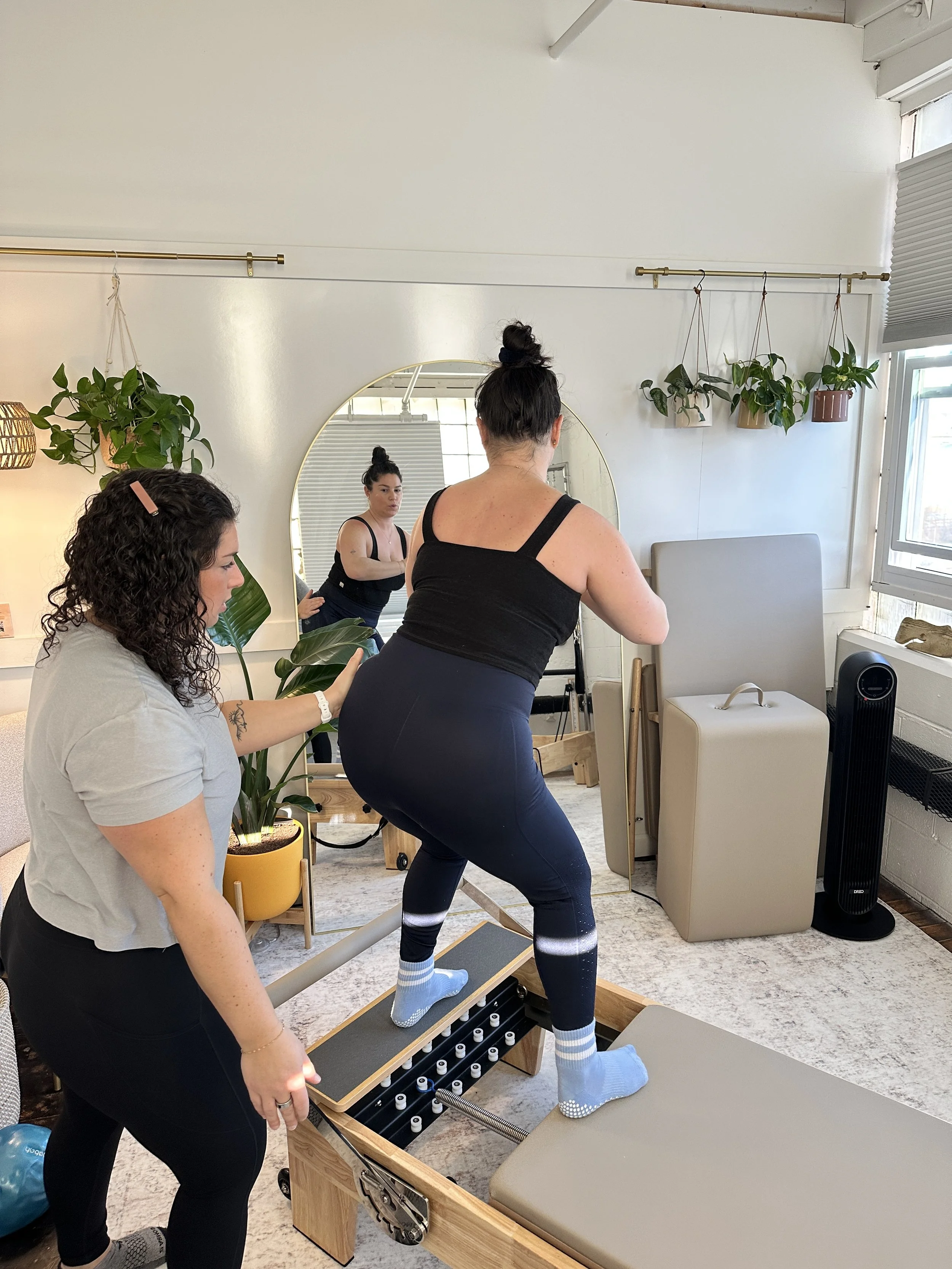 A woman is practicing a fitness exercise on a reformer machine assisted by a trainer in a well-lit room with plants, a mirror, and large windows.