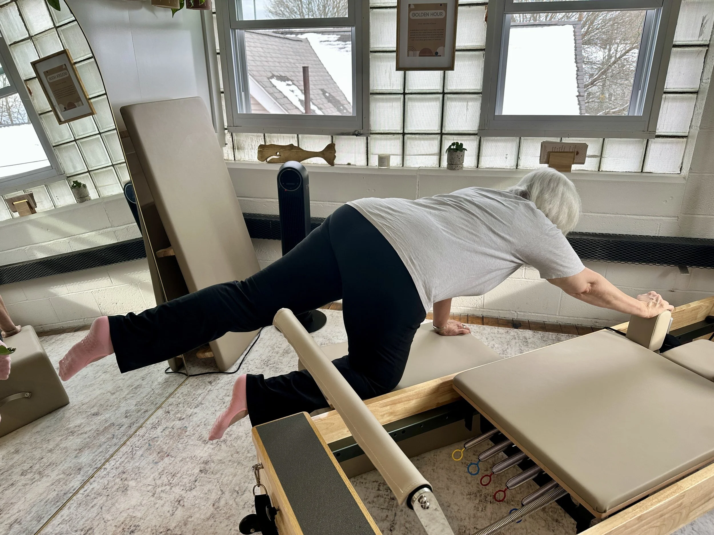 An elderly woman doing a stretch exercise on a specialized Pilates reformer machine in a bright room with large windows and a view of rooftops outside.