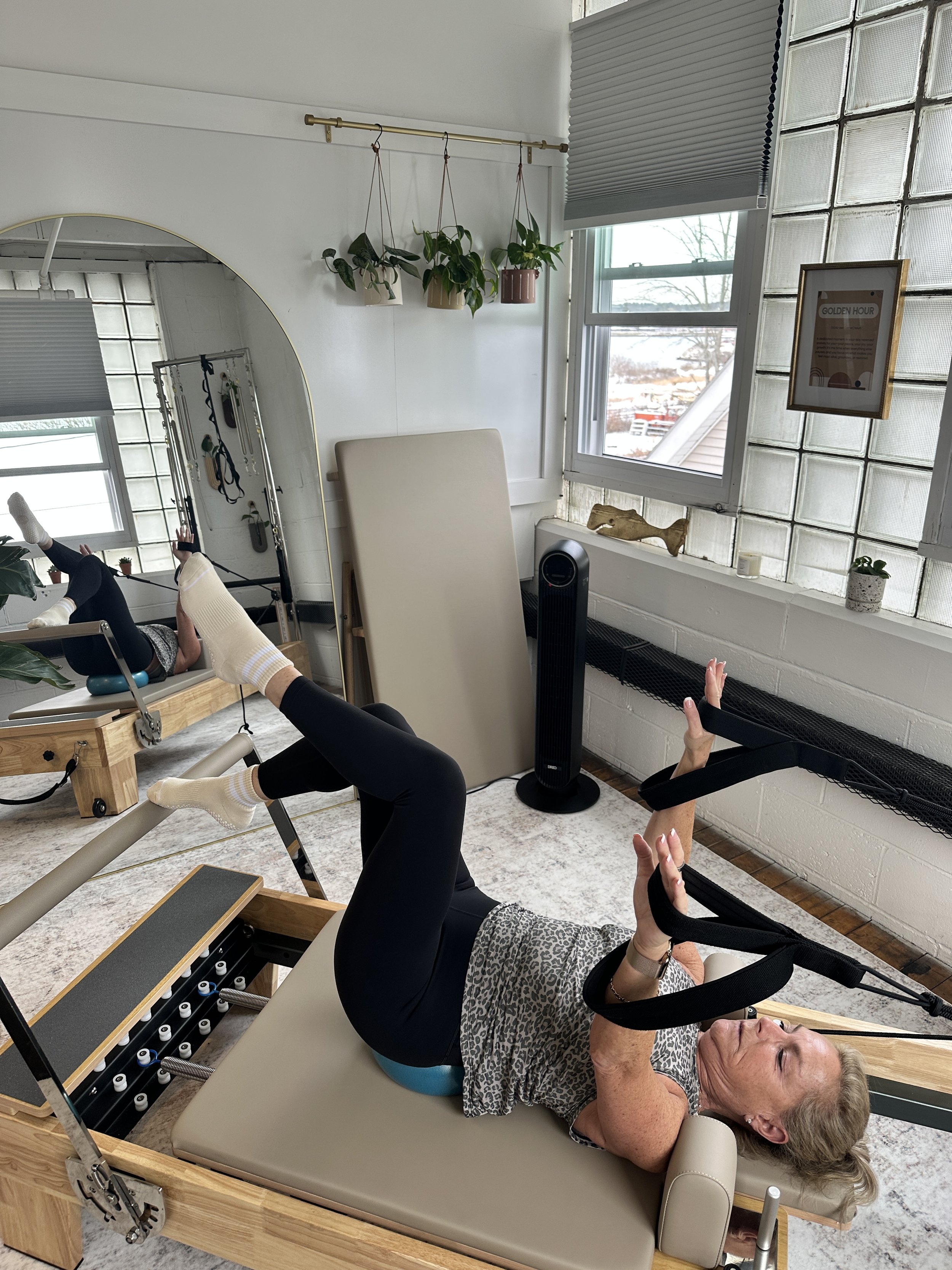 An elderly woman lying on a Pilates reformer machine, exercising with resistance bands in a bright, modern studio with large windows and plants.