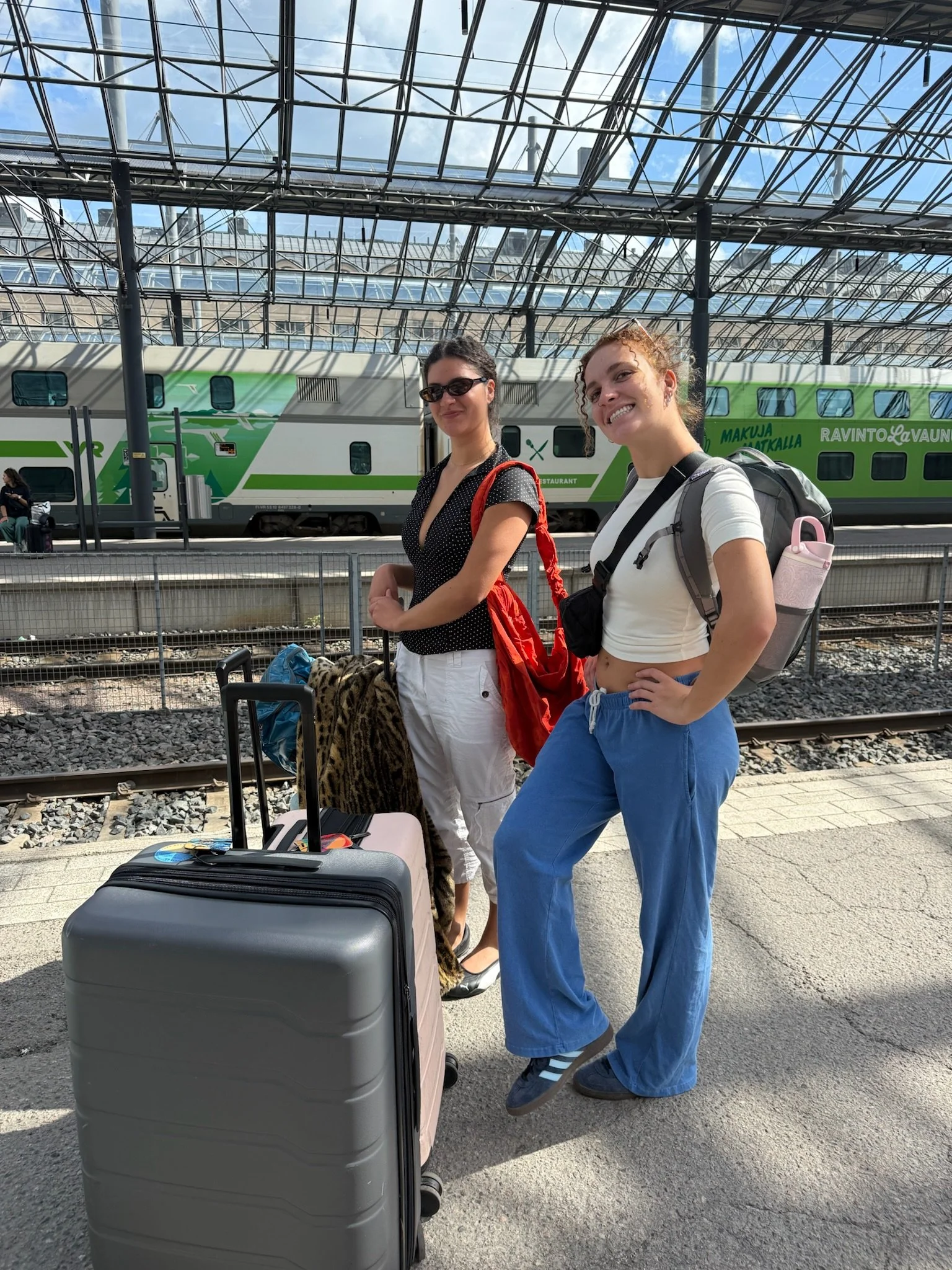 Two women standing at a train station with luggage in front of a green and white train under a large glass canopy.