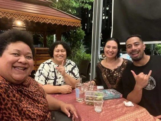 Four people sitting at a table outdoors, smiling for the camera. The table has glasses of water and some menus or books. The setting is lively with greenery and decorative lighting.