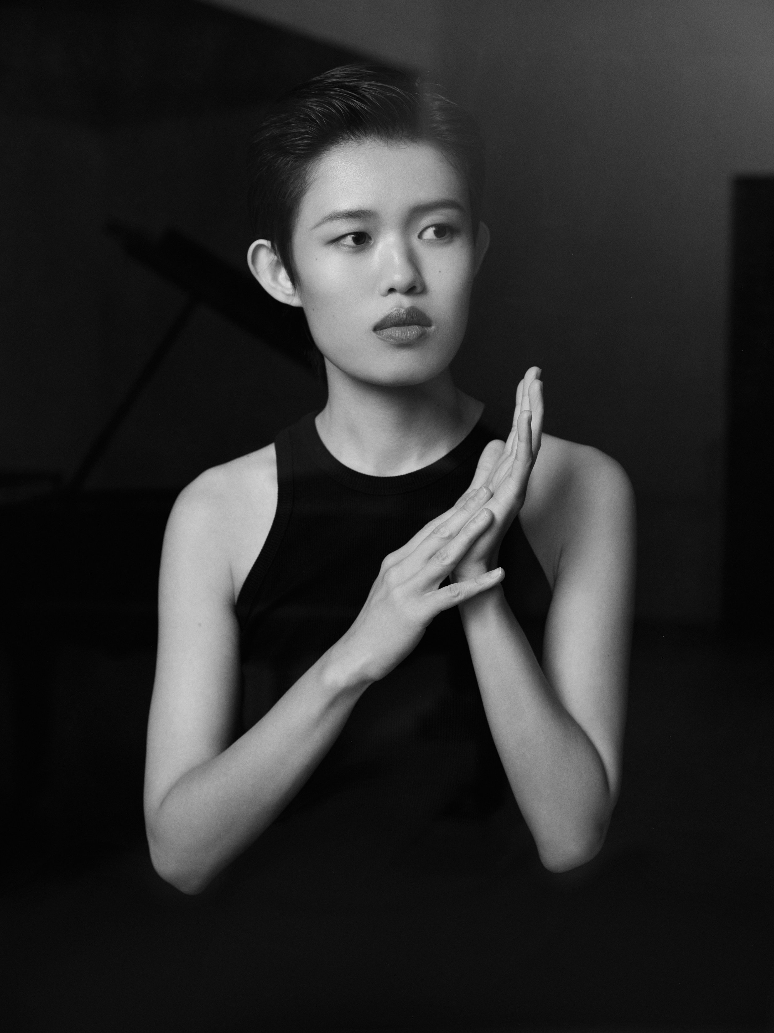 A young person with short hair wearing a sleeveless top, looking thoughtfully to the side, with their hands together near their face, in a black and white portrait.