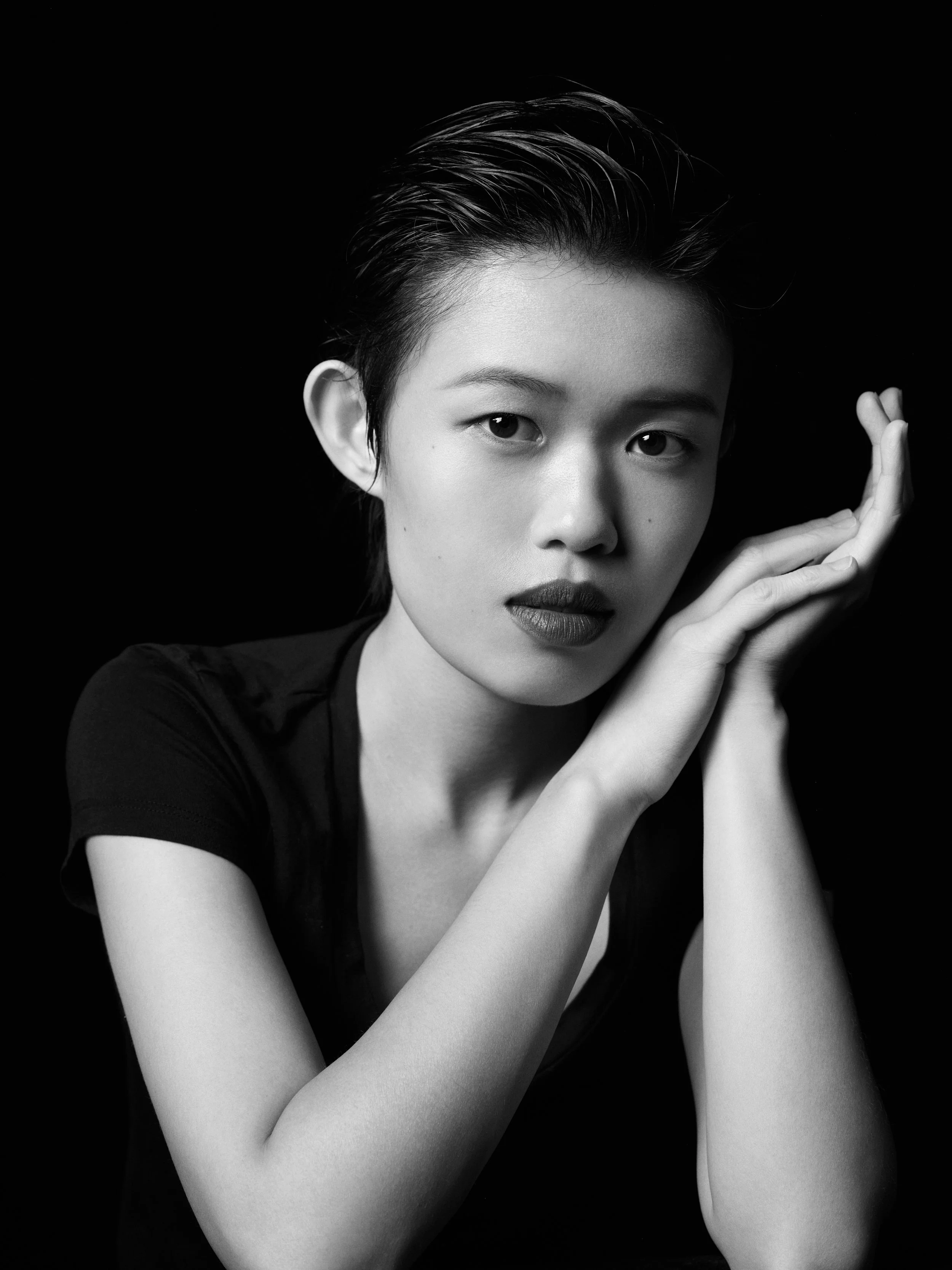 Black and white portrait of a person with short, wet hair, resting their chin on their hand, against a dark background.