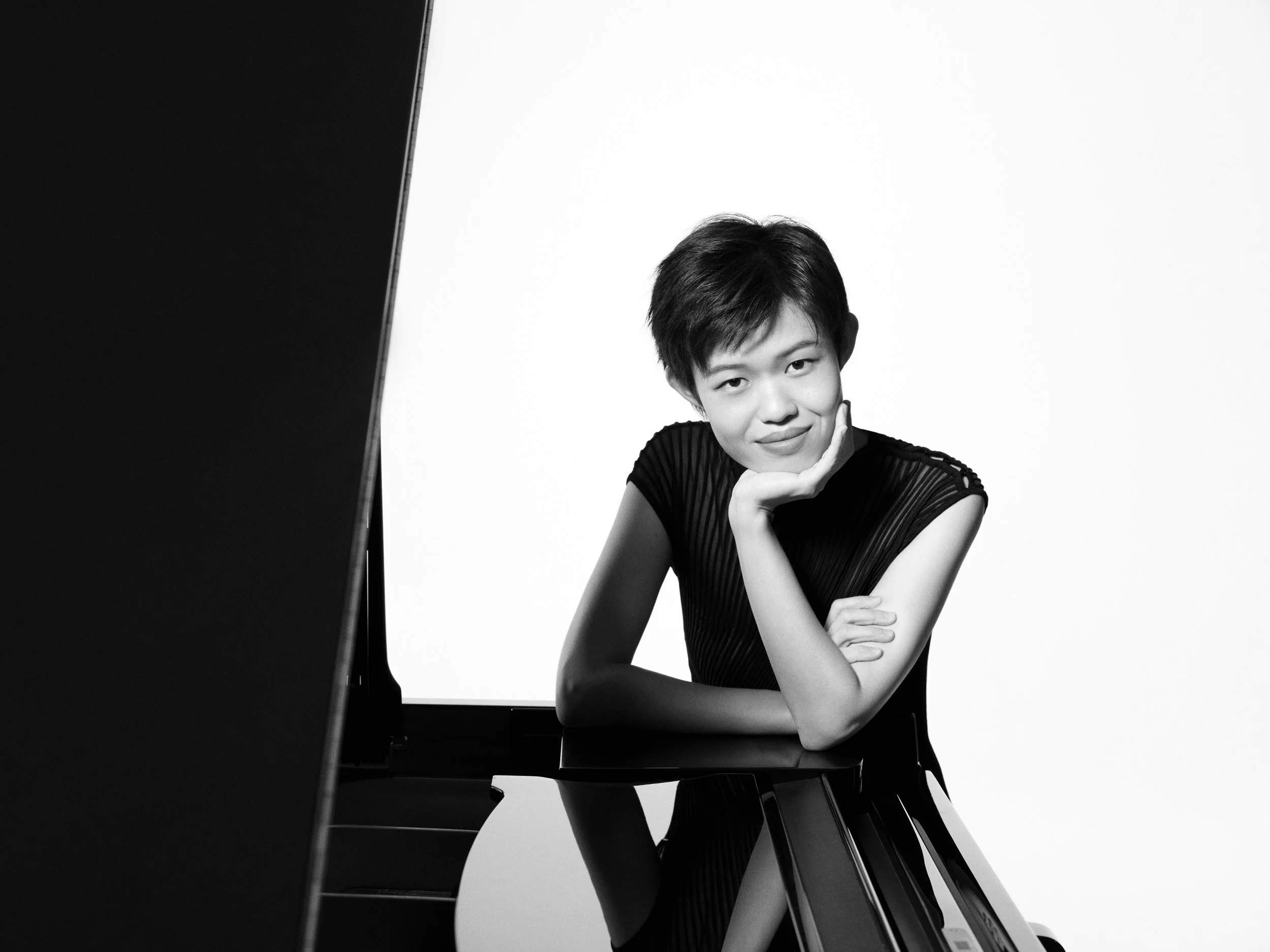 A black and white portrait of a person with short hair, sitting with their head resting on their hand, looking at the camera with a gentle smile, against a plain light background.