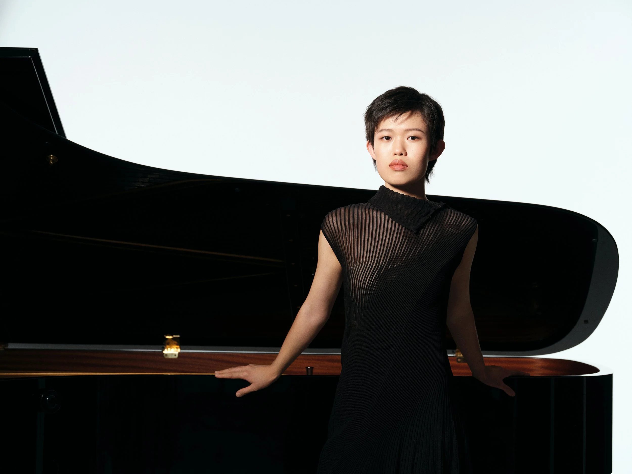 Young person in black dress standing beside a grand piano with white background.