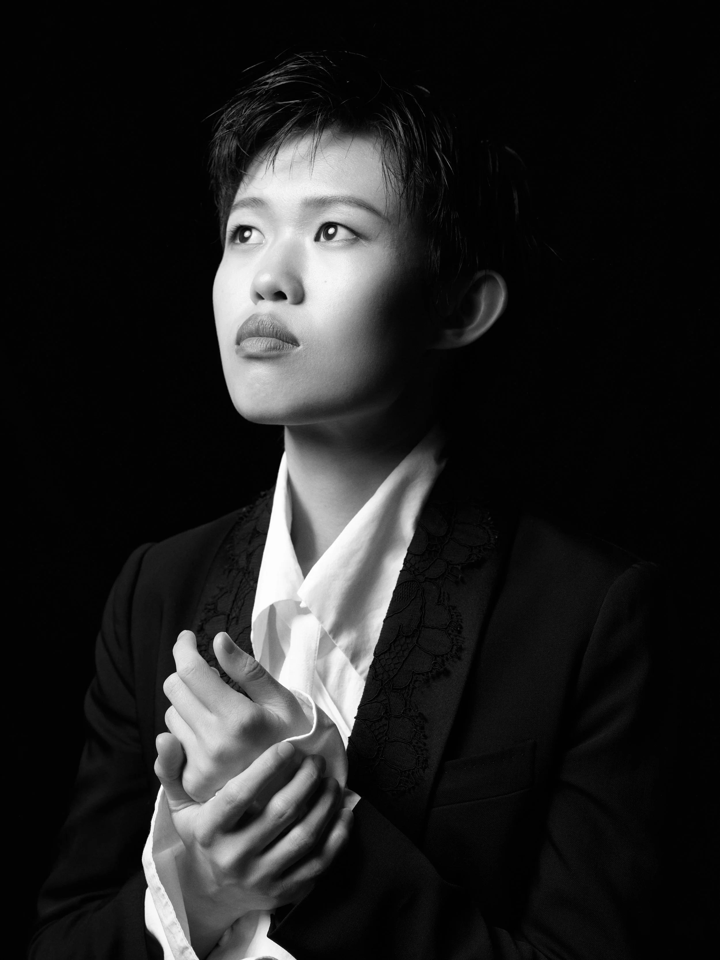 Black and white portrait of a person with short hair, looking upwards with a serious expression, wearing a white shirt and dark blazer with lace details, hand placed over their chest, against a dark background.