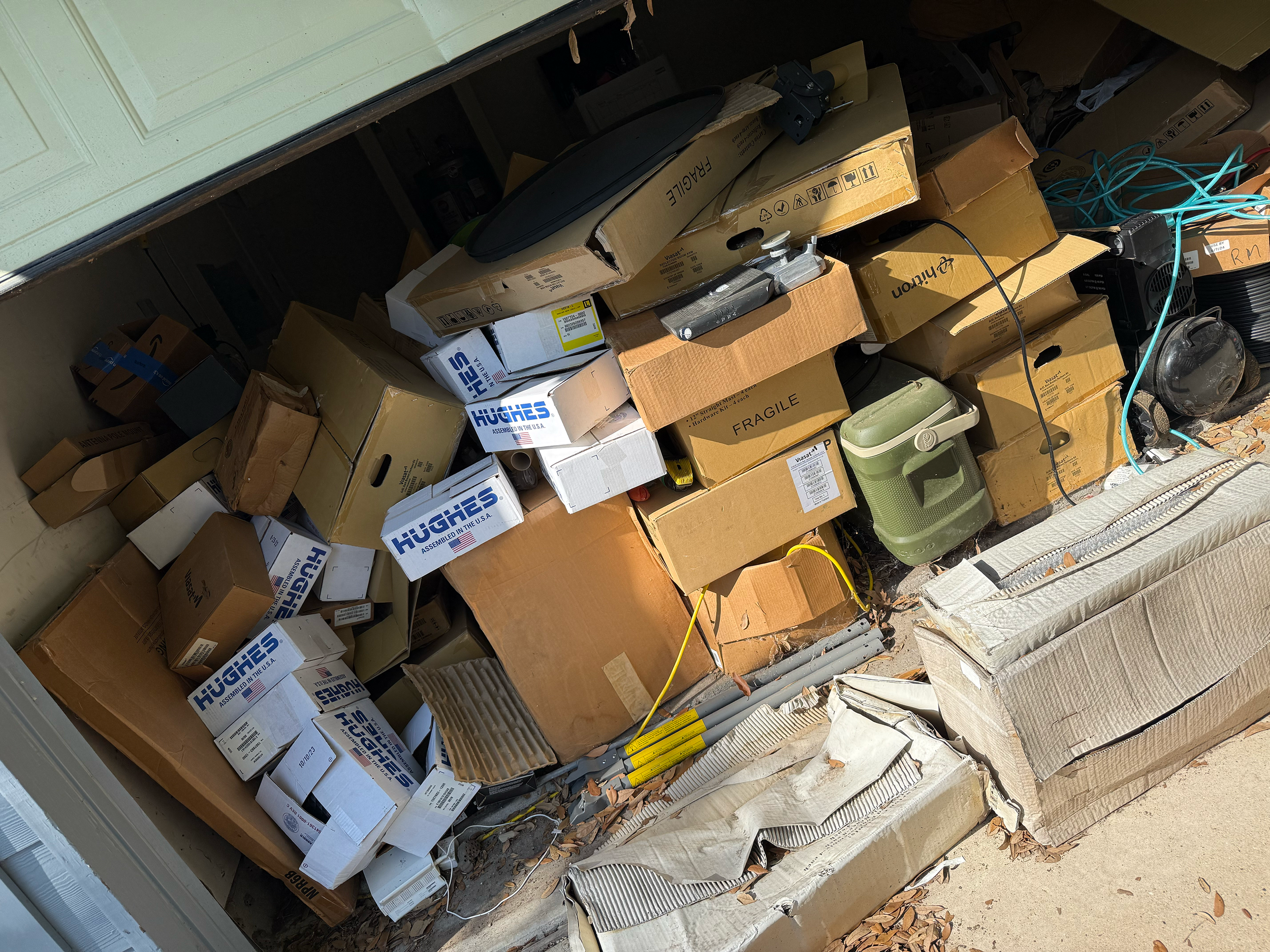 A cluttered garage with many cardboard boxes, some labeled 'fragile,' stacked haphazardly, with a green plastic container, electrical cords, and various miscellaneous items on the floor.