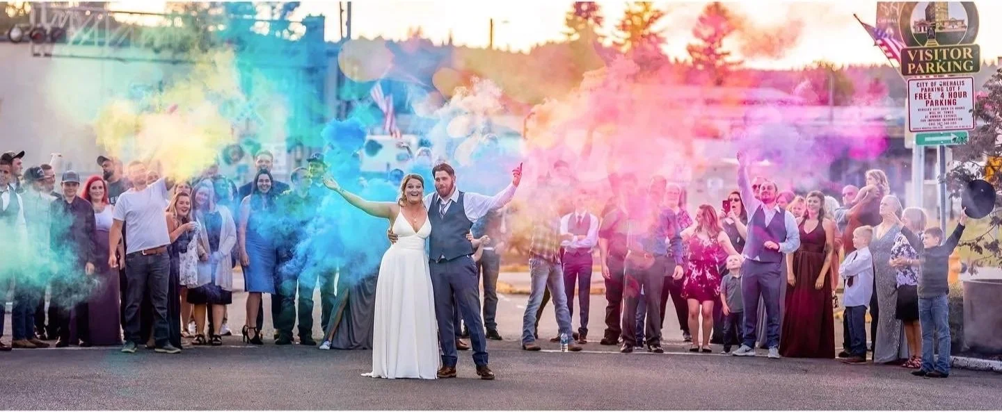A bride and groom celebrating in front of a crowd at a wedding, holding colorful smoke bombs during sunset.